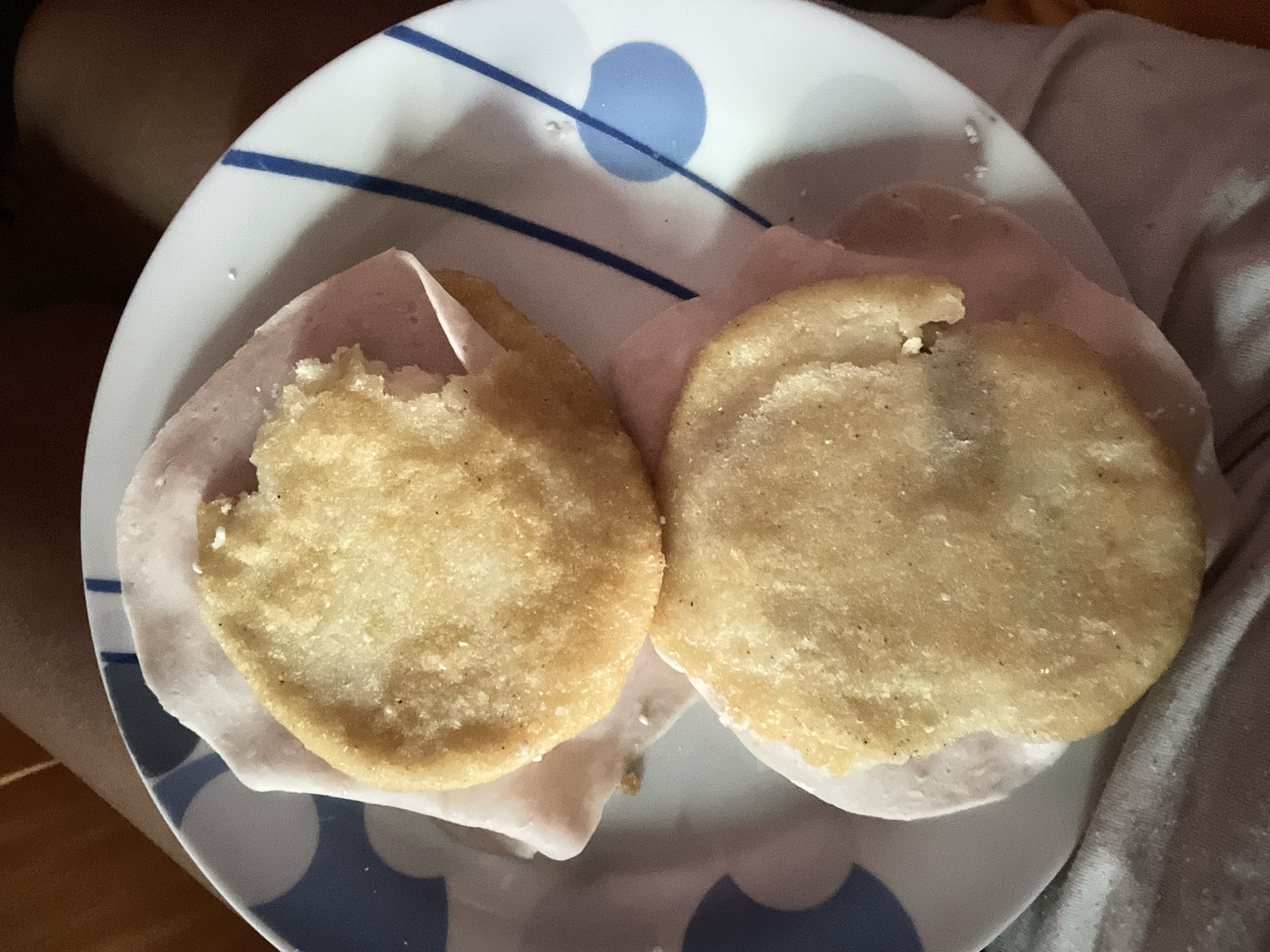On a white plate with blue circular and linear designs, two arepas are visible, each layered with a slice of ham. A bite has been taken out of the left arepa. The plate is positioned in Unknown, Unknown.