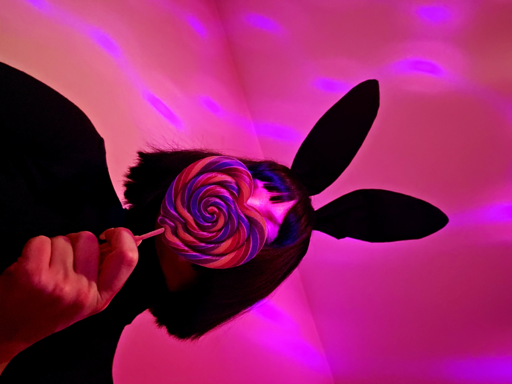 A person wearing black bunny ears holds a lollipop in front of their face. The background is lit with pink and magenta lighting. The location is not specified.
