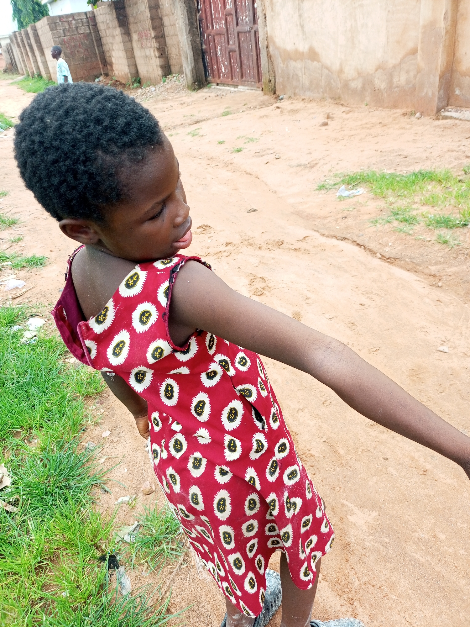A child with dark skin and black hair, wearing a red dress with a white and yellow floral pattern, stands in an outdoor setting with sandy ground and sparse green grass. The child is positioned in a three-quarter view, facing away from the camera and looking to the right. Their right arm is extended outwards. In the background, a brick wall and a wooden door are visible. A person in a light blue garment can be seen walking in the distance. The scene appears to be in a residential area in Tempure, Nigeria.