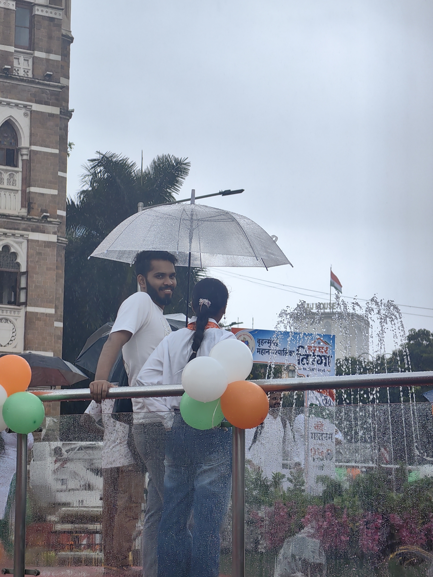 The image captures a street-level view during what appears to be a parade or public event in Mumbai, India. The scene is set against a backdrop featuring a tall, historic brick building with Gothic-style architecture, likely a landmark structure in the city. In the foreground, a clear umbrella is held overhead, indicating rainy weather, with water droplets visible on its surface and on other reflective elements in the scene.
Two individuals are prominent in the frame: a young man with a beard, wearing a white t-shirt, is smiling and looking towards the camera. Beside him, a young girl with her hair in a braid, wearing white clothing, is partially visible. They are both holding onto a metal railing, suggesting they are on an elevated platform or vehicle.
Several balloons, in orange, white, and green, are tied to the railing, adding a festive element to the scene, and are arranged in a manner reminiscent of the Indian flag's colors. In the background, a fountain sprays water, and a flag, also appearing to be the Indian flag, is visible flying from a pole. A banner with text in what seems to be Marathi script is also present. The overall environment suggests an outdoor public gathering in an urban setting, possibly during a national holiday or celebration, with monsoon rains contributing to the atmosphere. The presence of the historic building and the fountain, along with the general urban infrastructure visible, points to a location in a developed area of Mumbai.