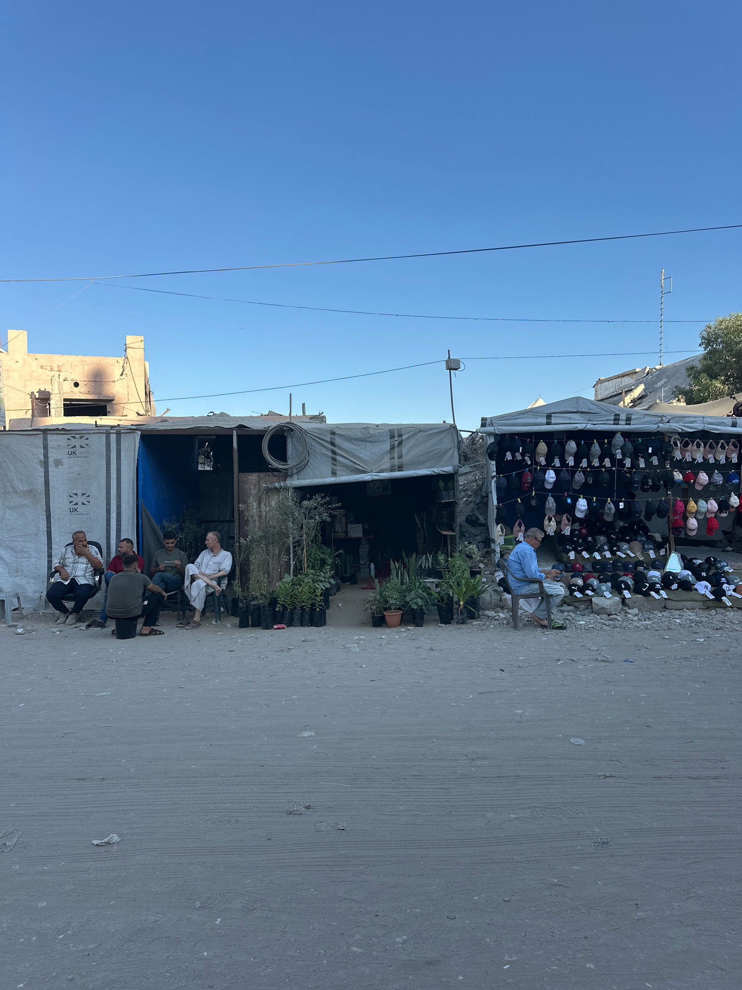 The image depicts a scene in the city of Gaza, Palestinian Territories, on a clear day. In the foreground, a dirt road shows tire tracks. In the background, several makeshift stalls are set up along a dusty street. On the left, a white tarp with "UK" logos is draped over a structure. Four men are seated on chairs outside this structure. One man is wearing a checkered shirt, another a red shirt, and two are in green shirts. A fifth man, in a white robe and white head covering, is seated on a chair facing them. Various potted plants are arranged in front of this stall.
To the right of this stall, another stall features a wide assortment of baseball caps displayed on a hanging rack and laid out on the ground. A man in a light blue shirt and light-colored pants is seated on a chair in front of this cap stall, looking down at something in his hands. The stalls appear to be made of corrugated metal and fabric awnings. In the far background, a damaged building with exposed concrete and dark openings is visible under a clear blue sky. Electrical wires are strung across the scene. The overall environment suggests an informal market or commercial area in Gaza.