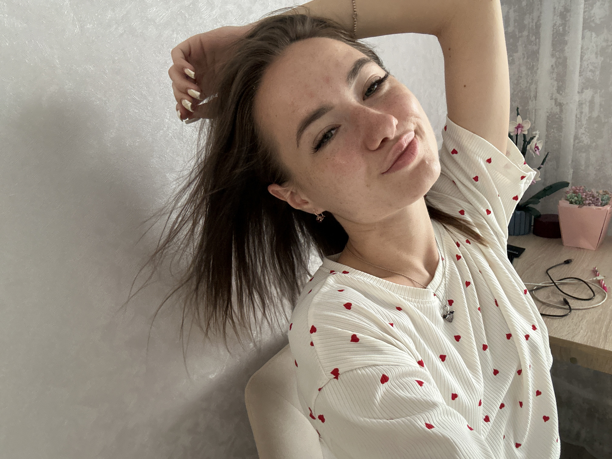 In an indoor setting, a young woman with dark hair and fair skin, adorned with a light-colored t-shirt featuring a pattern of small red hearts, poses with her right arm raised, hand resting on her head. Her left side is partially visible, and she is seated, with her hair cascading over her shoulder. The background consists of a textured, light-colored wall and a desk area with some items like potted plants and cables.