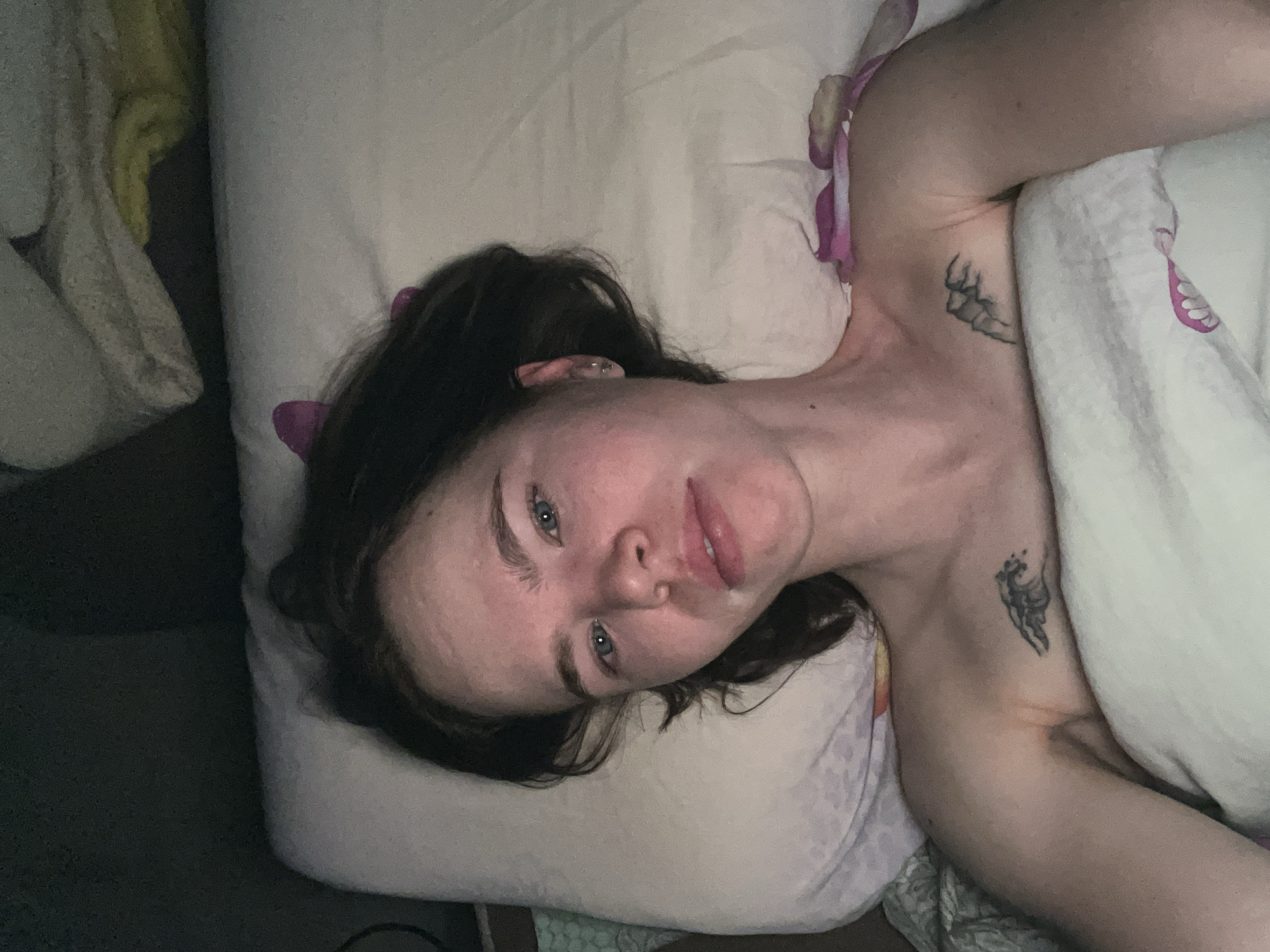 A woman is lying on a bed with a white pillow and a white blanket with pink accents. She has dark hair and blue eyes. There are two visible tattoos on her left shoulder and upper chest. The background includes a dark area, potentially a wall or another part of the room. There is no evidence to confirm the location of the image being in Chaikovsky, Russia.