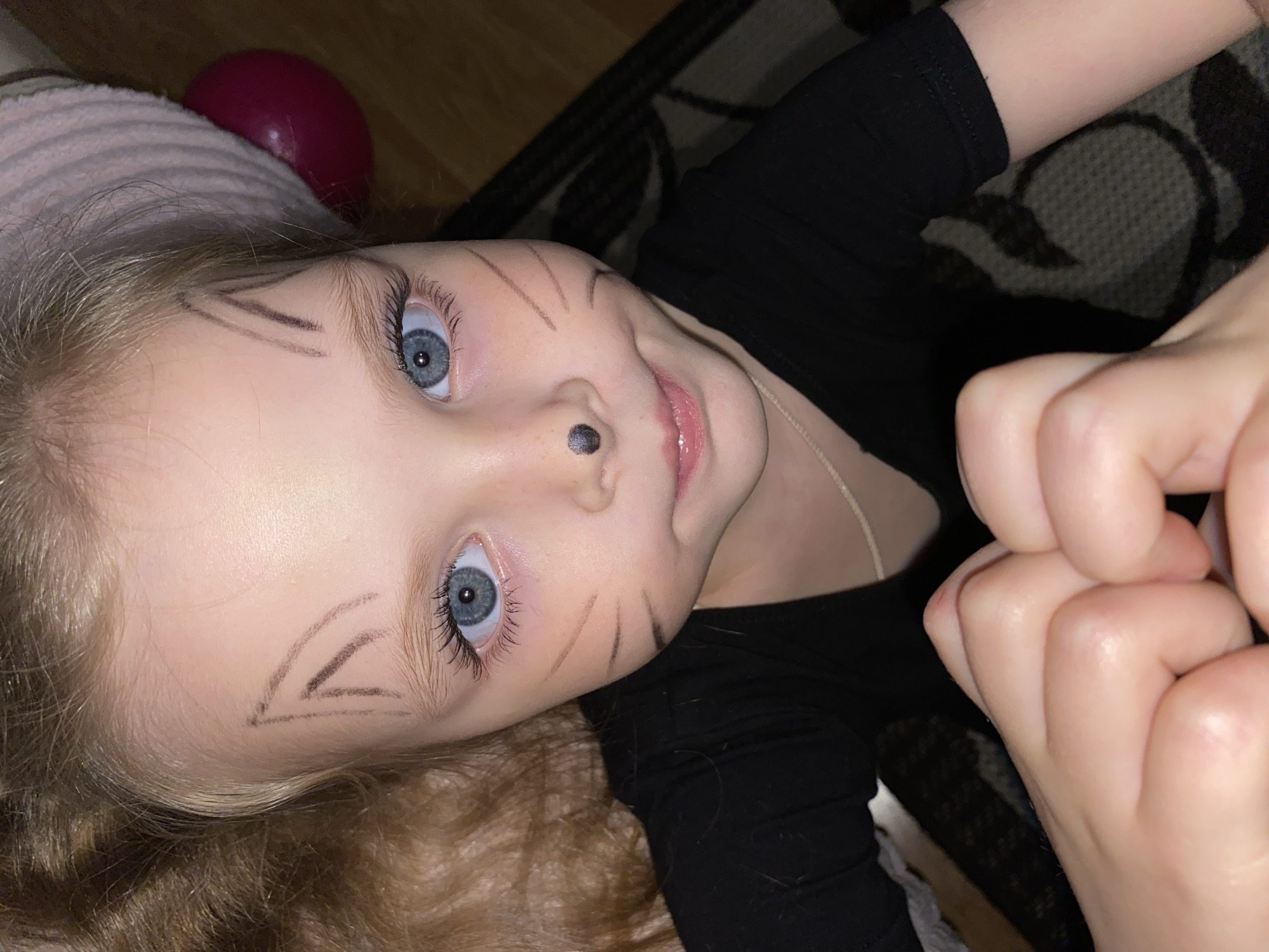 This tilted portrait captures a young child lying down, looking directly upwards with bright blue eyes and a soft, engaging smile. The child has light blonde or light brown curly hair and wears a black long-sleeved shirt. Their face is adorned with playful cat-like face paint, featuring drawn whiskers on both cheeks and a small black dot on the nose to mimic a cat's nose.
On the left side of the frame, behind the child's head, a pink textured blanket or pillow is visible, along with a partially obscured dark red spherical object, possibly a toy or ball. The child's hands are visible on the right, gently interlocked. The setting is indoors, featuring a patterned dark rug or carpet and what appears to be a wooden floor. The artificial lighting suggests the photograph was taken in the evening or at night. The child's expression conveys a sense of innocence and playfulness, likely engaged in a dress-up activity. There is no visible text in the image. The location context for this scene is Votkinsk, Russia.