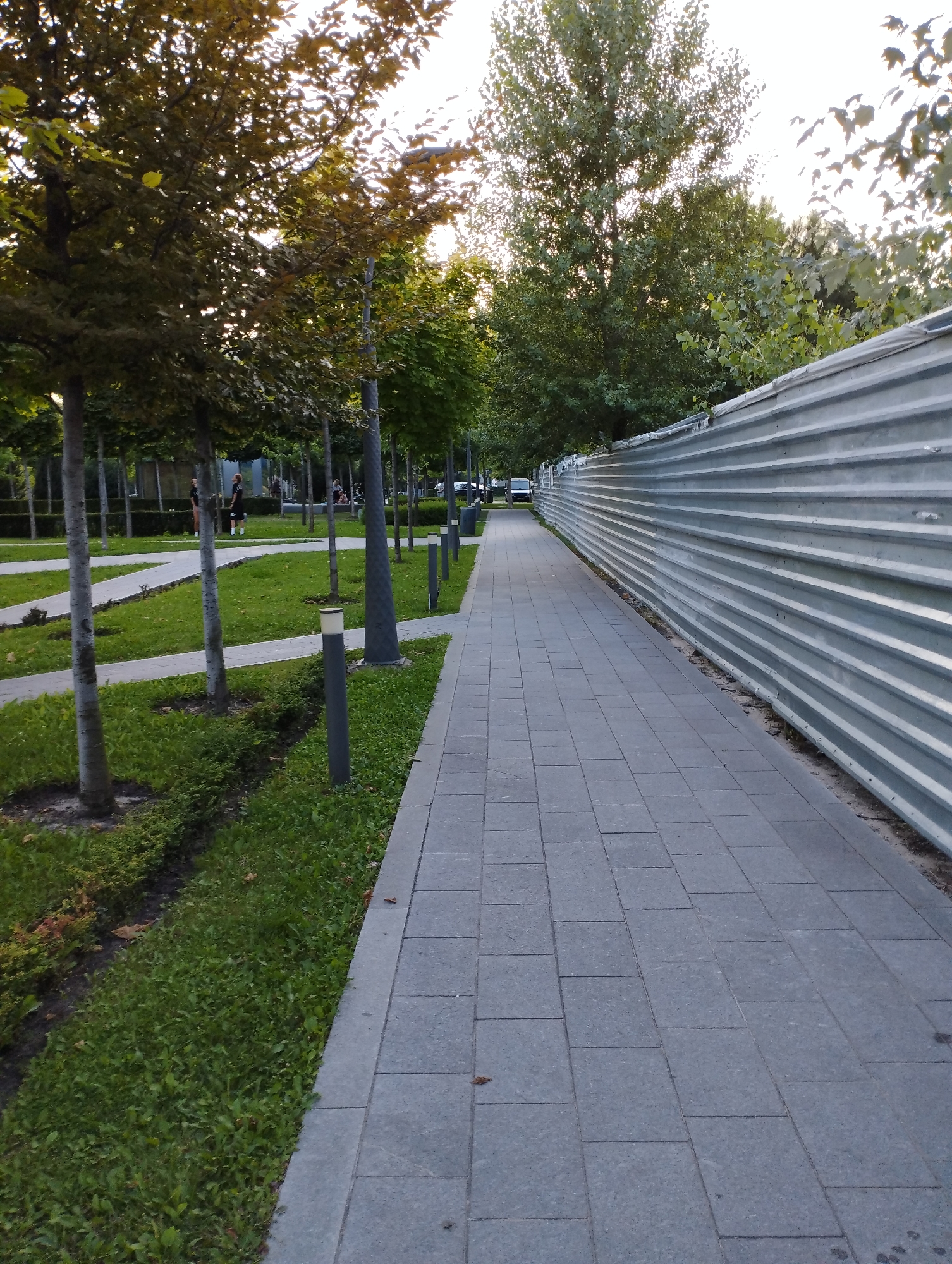 The image depicts a paved walkway running alongside a corrugated metal fence. The walkway is composed of gray square tiles. To the left of the walkway is a grassy area with several young trees planted in rows. Some of these trees have visible light fixtures at their bases. In the distance, on the left side of the image, there are several people walking and standing, some near other trees and a white path. The fence on the right extends into the distance and appears to be a temporary barrier. The overall environment suggests an outdoor public space, possibly a park or a newly developed area, with a mix of greenery and constructed elements.