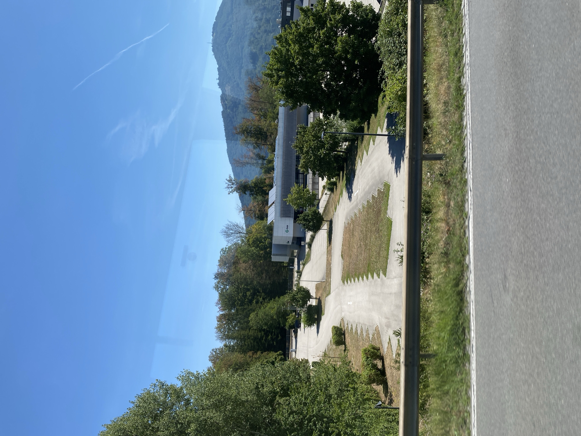 The image depicts an exterior scene on a bright, sunny day, likely in the vicinity of Gmunden, Austria, given the presence of a modern building with a logo that reads "Sax" and solar panels on its roof. The foreground features a portion of an asphalt road with a white line, suggesting a roadside view, possibly from a vehicle. A metal guardrail separates the road from a grassy embankment and a large, paved parking lot. The parking lot is mostly empty, with a few strategically placed trees and shrubs.
Behind the parking lot, a modern, rectangular building stands with a light-colored facade. The "Sax" logo is prominently displayed on the building. Beyond the building, a dense line of trees forms a natural backdrop, leading up to a large, green, tree-covered mountain in the distance. The sky is a clear, bright blue with a few wispy white clouds and contrails. The overall impression is one of a commercial or industrial area nestled within a scenic, natural landscape. There are no people visible in the image.
