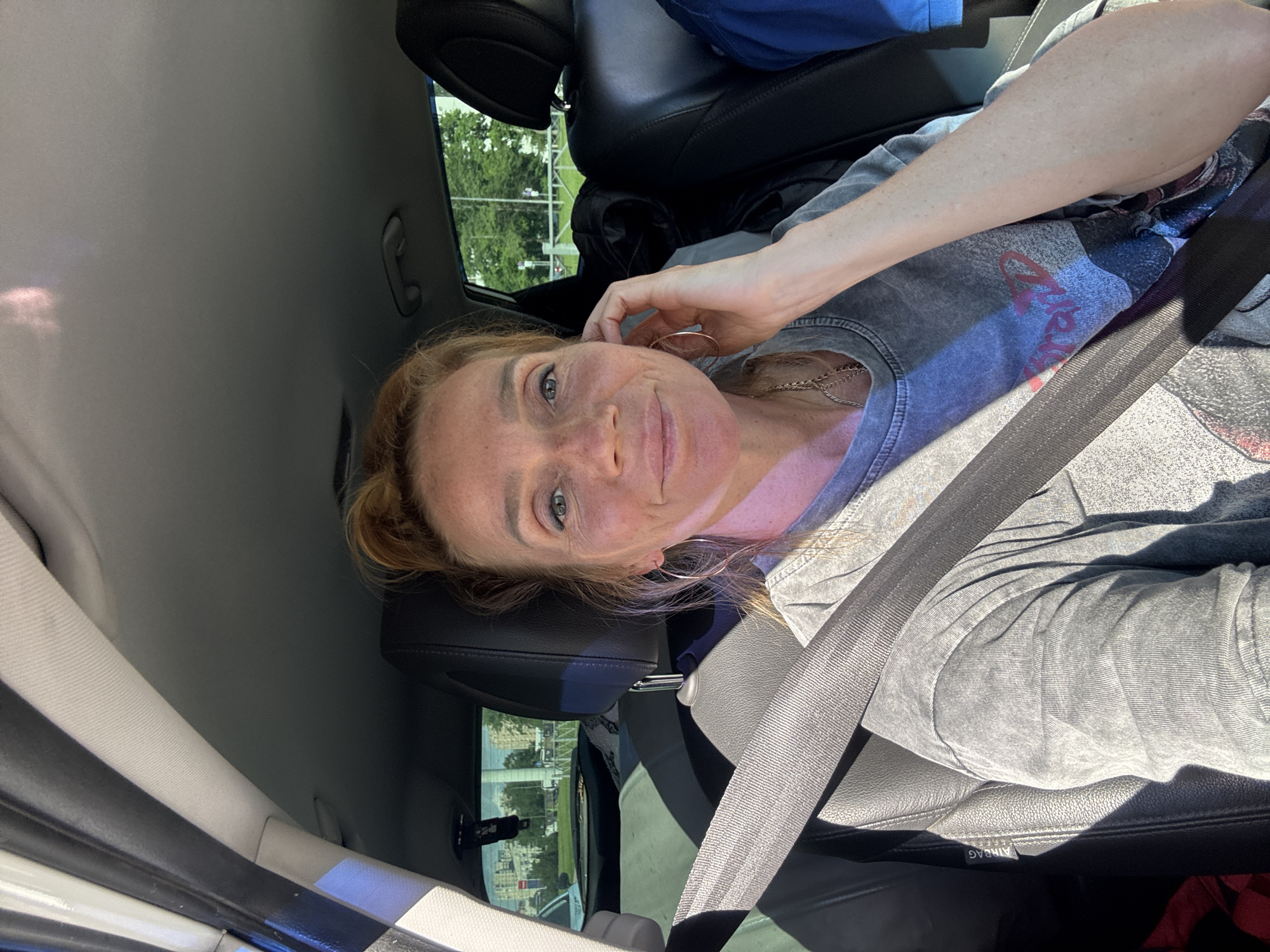 A female individual is seated in the front passenger seat of a vehicle, secured by a dark grey seatbelt. She has fair skin with visible freckles across her nose and cheeks, blue eyes, and reddish-brown hair styled in an updo. She is wearing a faded grey, short-sleeved t-shirt with a partially visible graphic design featuring red lettering and an image. Large hoop earrings are worn, along with a delicate chain necklace. Her left hand is raised towards her face.
The interior of the vehicle features black leather or synthetic material seats, with an "AIRBAG" label visible on the side of the passenger seat. The headliner is light grey, and a grey grab handle is present above the passenger door. An object hangs from the rearview mirror.
Through the windows, an urban or suburban environment is visible. On the driver's side, buildings, a white car, and green foliage are discernible. On the passenger's side, beyond a white fence or barrier, numerous green trees and distant structures are apparent, suggesting a roadside or park area. Strong sunlight casts distinct shadows within the vehicle. This scene takes place in the city of Yukki, Russia.