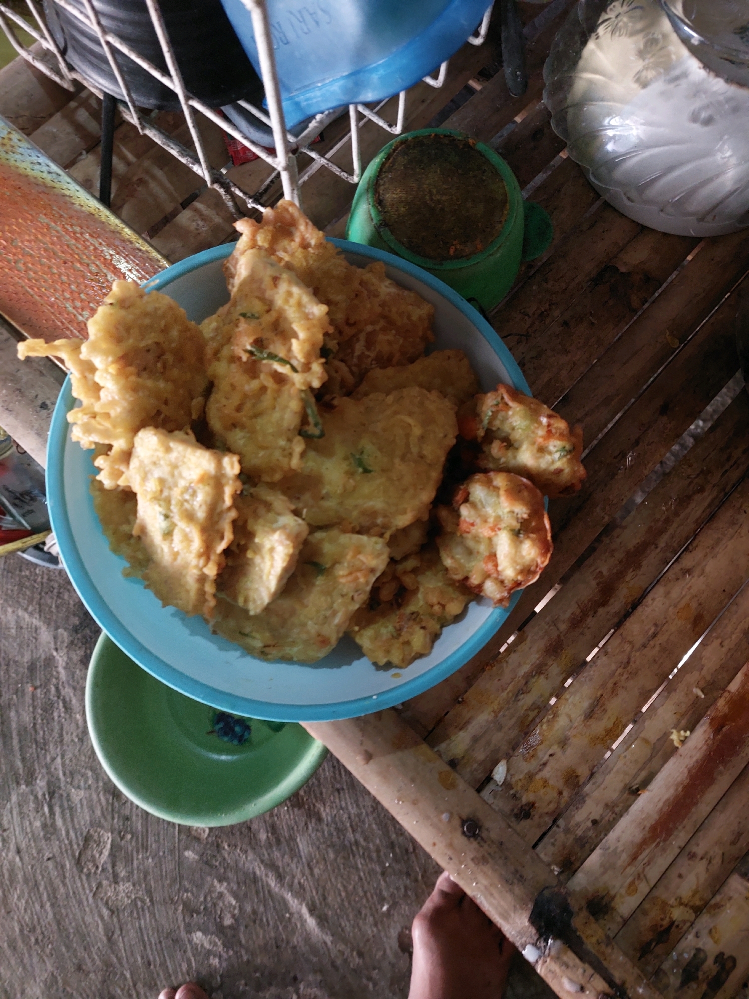 The image shows a close-up, high-angle shot of a light blue bowl filled with fried food, possibly bakwan, placed on a bamboo table. The fried food items are golden brown and appear to be made with vegetables. Underneath the blue bowl is a green bowl. The background includes a white wire rack with a blue container, a green object, and a clear plastic container with a lid. The surface the food is on is made of bamboo planks. The bottom left of the image shows a rough, greyish surface. The lighting is dim, casting shadows. The scene appears to be outdoors, given the natural materials and the informal setting. The presence of the word "SARANGAN" on the blue container suggests the location of the image is Sarangan, Indonesia, which is known for its lake and is a popular tourist destination.