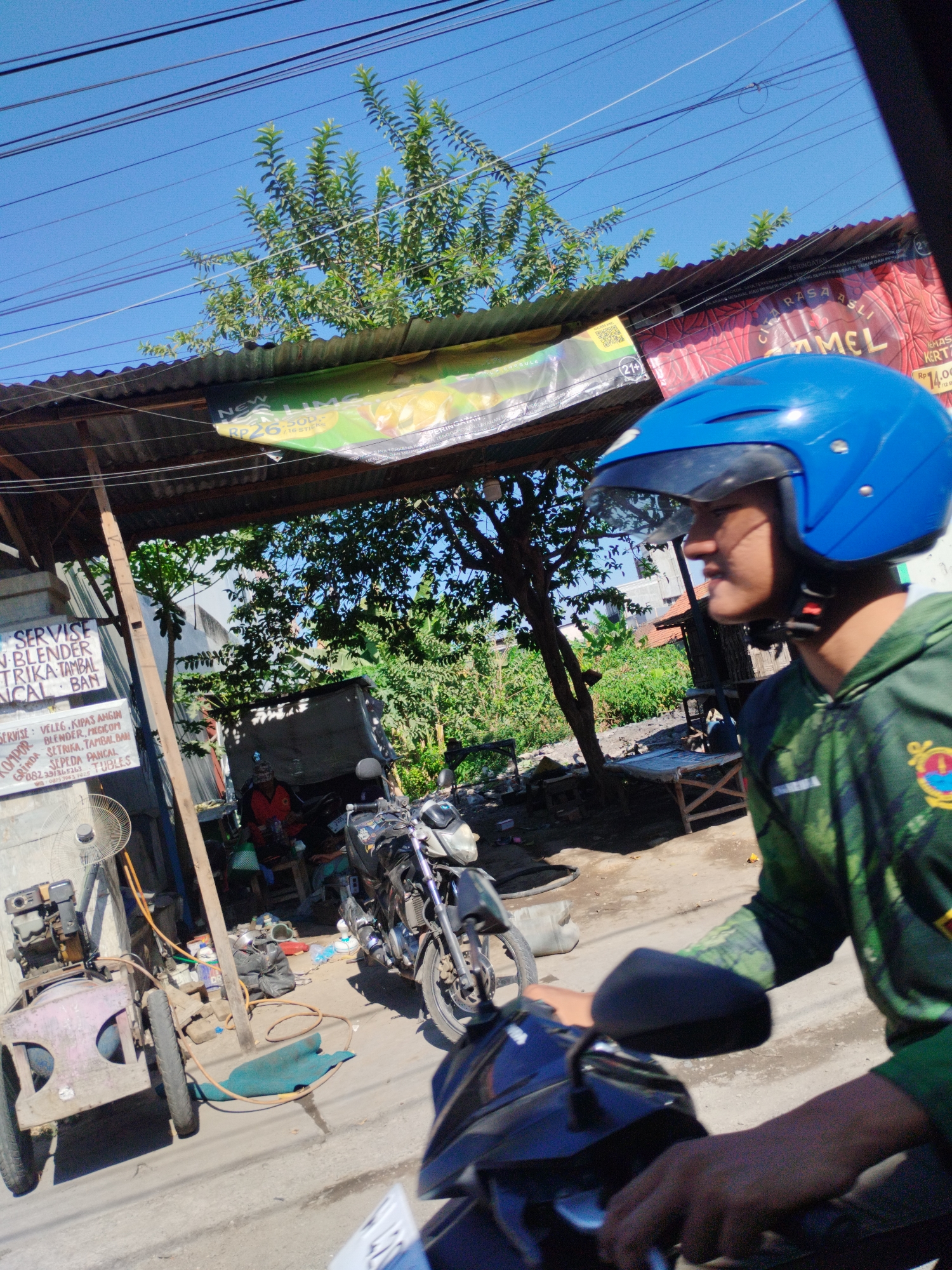 The image depicts a roadside scene in Surabaya, Indonesia, under a bright blue sky. A young man wearing a blue helmet and a green hooded jacket is riding a motorcycle, his right hand on the handlebars. In the foreground, the motorcycle's black rearview mirror is visible. The background reveals a small workshop or stall with corrugated metal roofing. Signs are posted on a wooden post, with one sign listing services such as "SERVISE: VELLIG, KUPAS ANGIN BLENDER, PENGOM SETRIKA, TAMBAL BAN, SEPEDA PANCHAL" and a phone number. There is a pink and grey air compressor with a fan attached on the left, and a black motorcycle is parked behind the air compressor. The ground is dirt and pavement. Further back, there is a person in an orange shirt sitting under a shelter, and trees with green foliage are visible. Advertisements are hung on the roofing, including one with the word "CAMEL" prominently displayed. The scene appears to be a typical street-side commercial area in Surabaya.