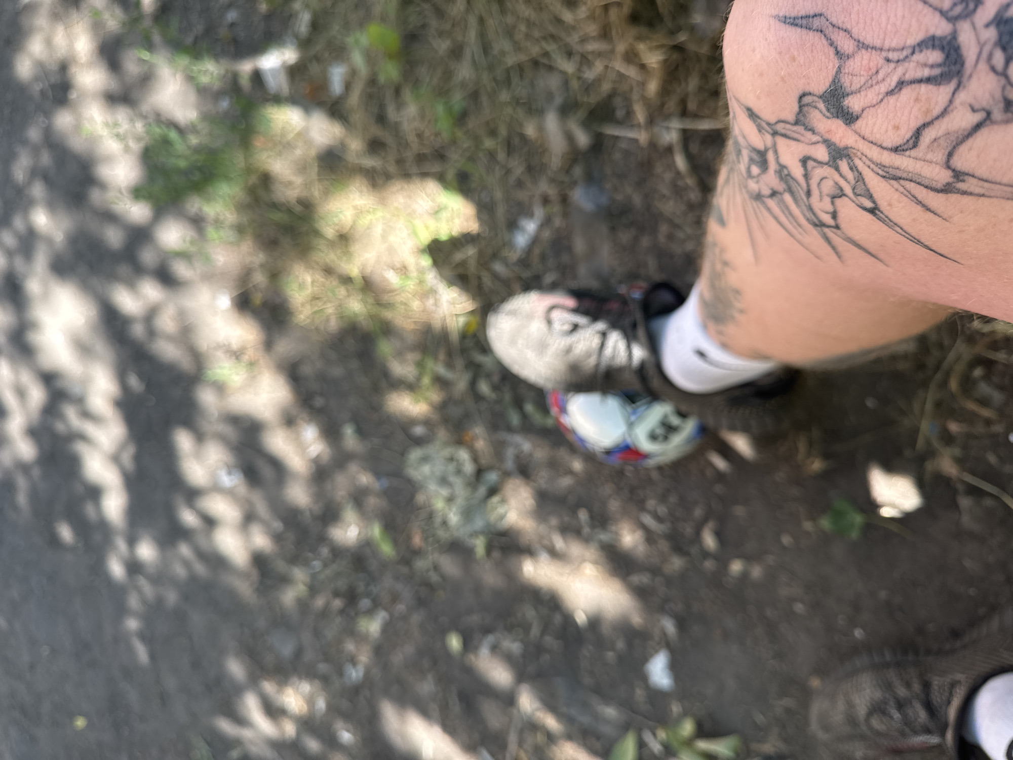 The image depicts a close-up view of a person's leg with a tattoo, a soccer ball, and footwear. The leg, which is tattooed with what appears to be a mythical creature or an abstract design, is in the foreground. A soccer ball, partially visible, is positioned near the person's foot, which is clad in a black and gray athletic shoe with white socks. The background is out of focus and appears to be an outdoor environment, possibly a park or a dusty area, with shadows from trees cast on the ground. The lighting suggests it is daytime.