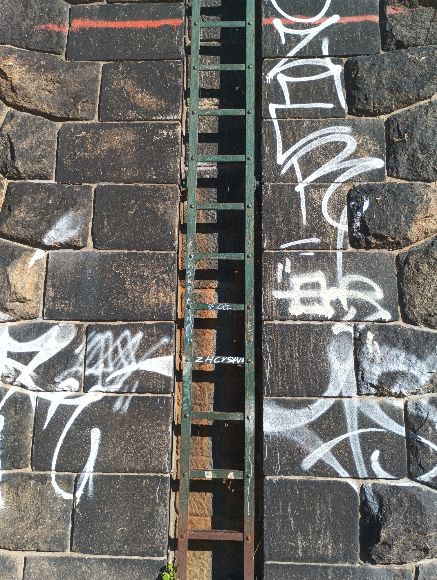 The image shows a close-up, eye-level, vertical shot of a stone wall with a green metal ladder attached. The stone wall is constructed from large, dark, rectangular blocks, some of which have rough, weathered surfaces. Graffiti in white spray paint is visible on the stone blocks on both the left and right sides of the ladder. A thin red line is painted horizontally across the top section of the stone wall on the left. The ladder, which appears to be made of rusted metal, is positioned in the center of the image. The rungs of the ladder are clearly visible, and the overall structure is vertical. Some of the stone blocks show signs of wear and tear, with dirt and debris accumulated in the crevices. The lighting suggests it is daytime, with sunlight casting shadows from the ladder onto the wall. The image appears to be taken in an urban environment, likely in Pilsen, Czechia, given the context provided. No people or animals are visible in the frame.
