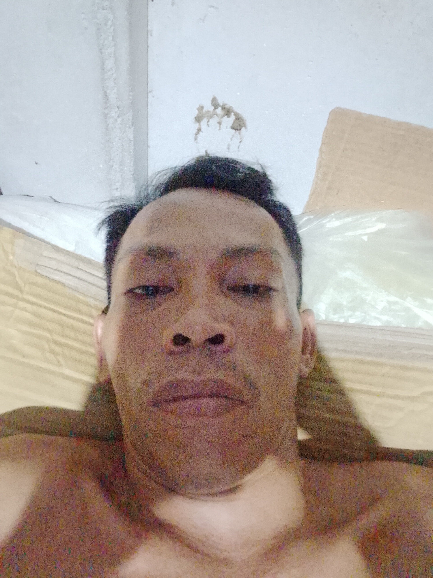 A close-up selfie shows an Asian man from the chest up. He has dark hair, a medium complexion, and is not wearing a shirt. Behind him are what appear to be blankets or a bed and a wall that shows signs of moisture damage. The location may be Bukit Ketok, Indonesia.