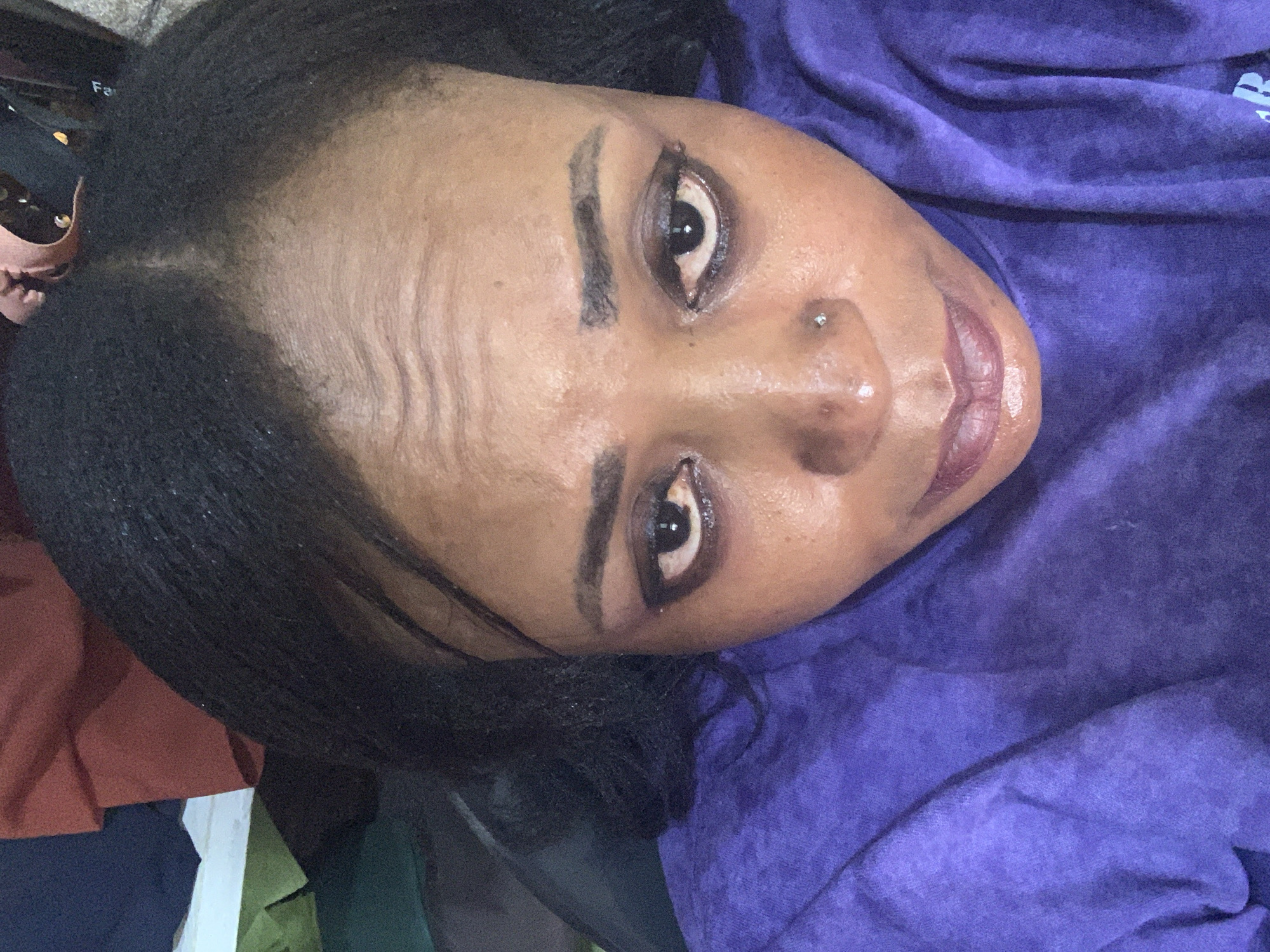 A close-up, overhead shot shows a Black woman's face from the forehead down to her upper chest, looking upward. Her dark hair is pulled back. She has light brown skin, dark eyebrows, and dark eyeshadow. She wears a nose ring and a purple shirt. The background includes unfocused reddish, green, and dark fabric. The image was taken in Sabon Tasha, Nigeria.
