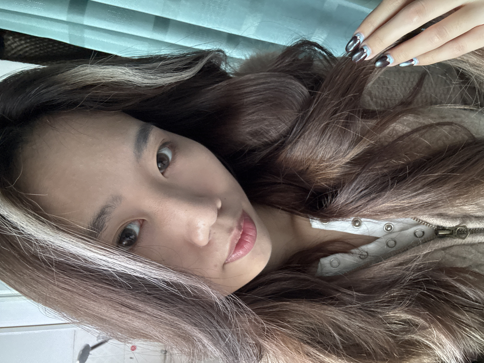 A female individual with long, wavy dark brown hair, featuring prominent white-blonde and lighter brown streaks near the front, is depicted in a close-up, frontal portrait. She possesses light brown eyes and is looking directly at the camera. Her lips are a light pink, slightly parted. She is wearing a light-colored, possibly white, Henley-style top with snap buttons, underneath a partially unzipped beige or light brown textured sweater or jacket. Her left hand is visible on the right side of the frame, gently touching her hair. The fingernails are manicured with a dark, glossy polish, accented by light blue crescent shapes at the base and small metallic studs.
The background indicates an indoor setting. To the left, a white wall and a partial view of a metallic fixture, possibly a faucet or showerhead, are discernable. To the right, a pleated light blue curtain hangs vertically. The lighting appears soft and uniform. The image is situated in an unspecified indoor location within Nan'an District, China.