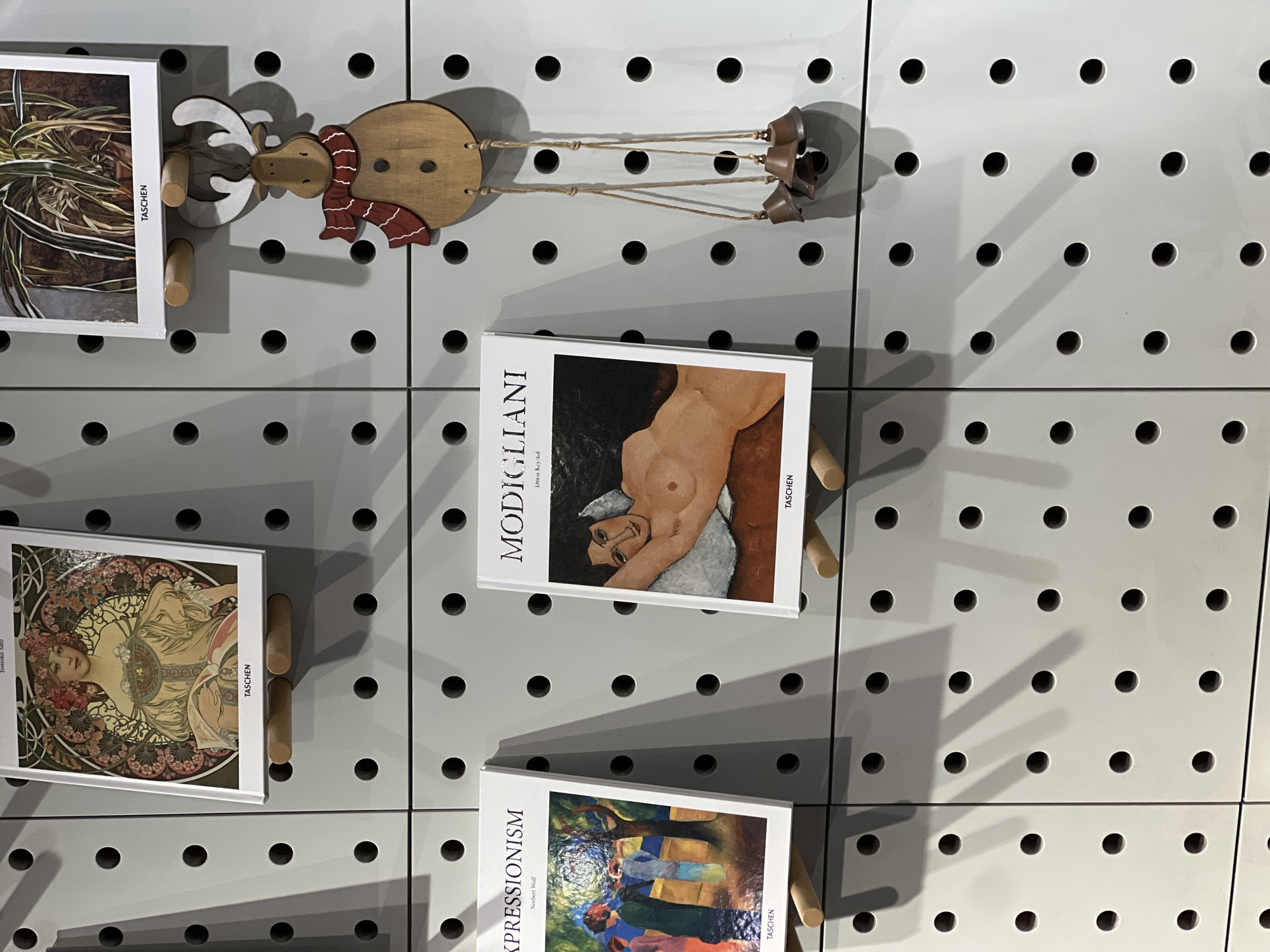 The image shows a display of TASCHEN art books and a wooden reindeer ornament on a gray pegboard wall. The books feature reproductions of artwork, including Modigliani paintings and other artistic styles. The books are held in place with wooden pegs inserted into the pegboard. The reindeer ornament consists of a wooden body and head with a red scarf, attached to metal bells by strings. The scene appears to be indoors, likely in a bookstore or gallery, although there is no information about being located in the city of Yerevan, Armenia.