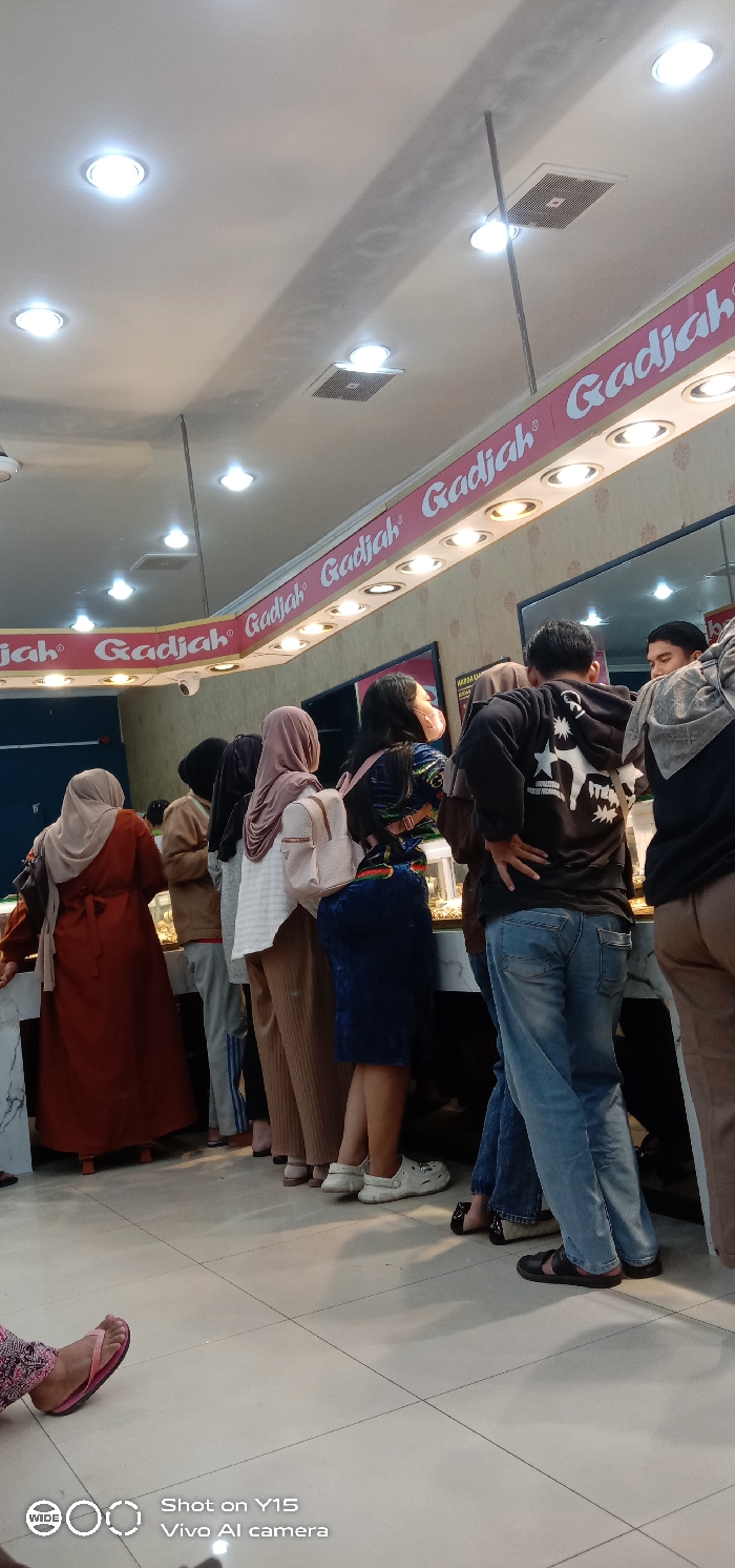The image displays an indoor scene within a commercial establishment in Probolinggo, Indonesia, identified by a prominent red banner with white lettering repeating "Gadjah," which illuminates the area. Approximately ten to twelve individuals, predominantly customers, are visible, mostly standing and oriented towards a long, white marble-topped counter featuring glass display cases containing various items.
On the far left, a woman wearing a rust-colored long dress and a light-colored hijab leans on the counter. Near the center, a man in a black hoodie with a white star graphic on the back and blue jeans stands with his hands on his hips, facing the counter. To his left, a woman in a blue velvet-patterned top and skirt, wearing white Crocs and a light-colored backpack, also faces the counter. Another woman nearby wears a light brown hijab, a white striped shirt, and brown pants, carrying a light-colored backpack. A male employee is partially visible behind the counter on the far right, appearing to interact with customers. One woman in the queue wears a light pink face mask.
The ceiling is white and fitted with multiple recessed circular lights and several square ventilation grilles. A small white CCTV camera is mounted below the "Gadjah" sign. The walls feature a light, textured pattern, with a section of dark blue wall visible on the left. The floor is composed of light-colored square tiles. A "Shot on Y15 Vivo AI camera" watermark is present in the bottom left corner. The scene depicts a busy retail environment, likely a pawn shop or a gold shop.
