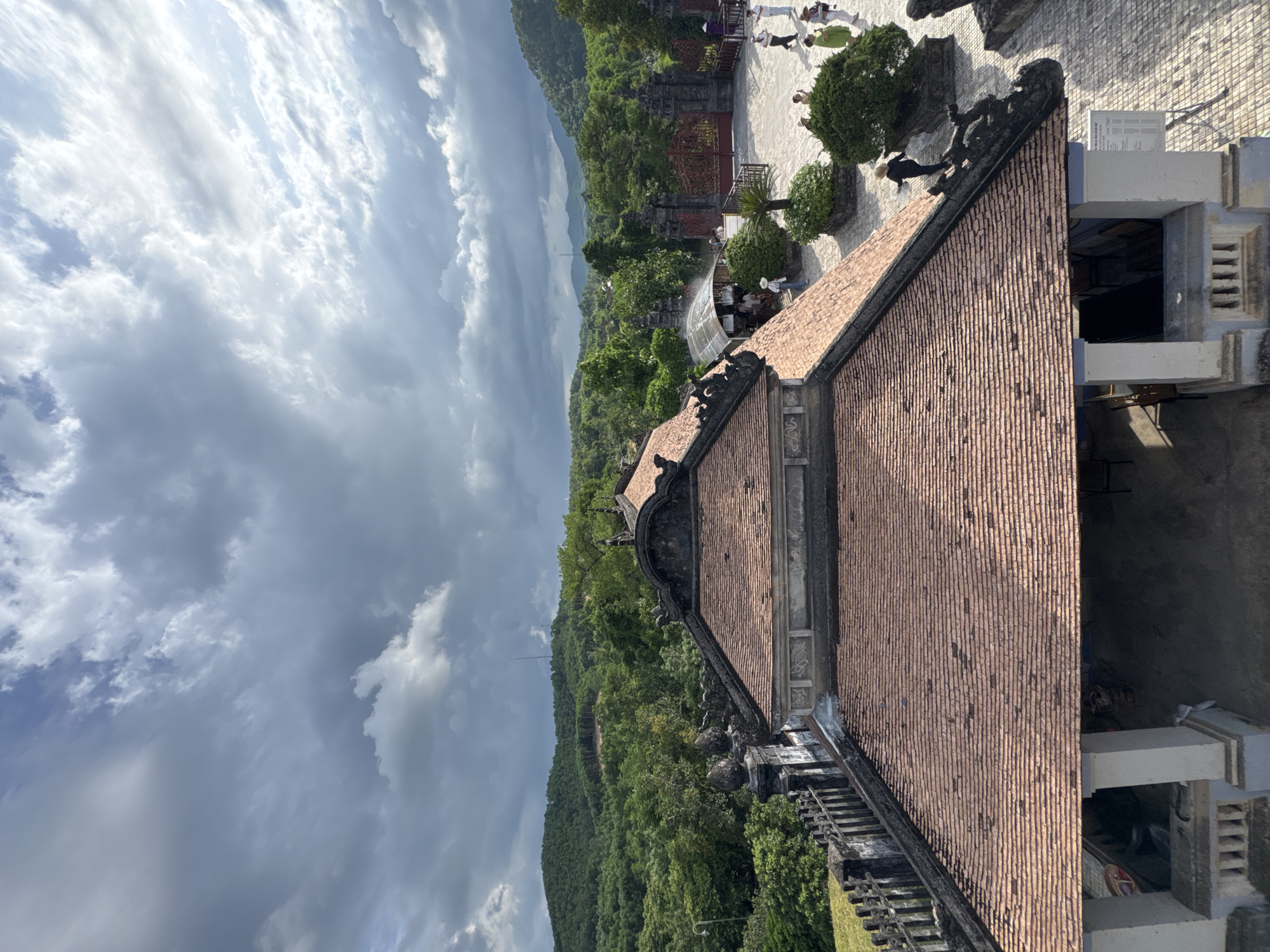 The image captures an overhead view of a traditional tiled roof building in Thuy Xuan Ward, Vietnam. The building features ornate dark grey decorative elements along its eaves and gables. Beyond the building, a courtyard paved with stone is visible, with several individuals walking and interacting. To the left of the courtyard, a dark fence with decorative posts runs along the edge of the property. A red gate stands prominently in the background, leading further into the complex. The scene is set against a backdrop of lush green rolling hills covered in dense trees, under a dramatic sky filled with dark, heavy clouds and patches of lighter, illuminated sky.