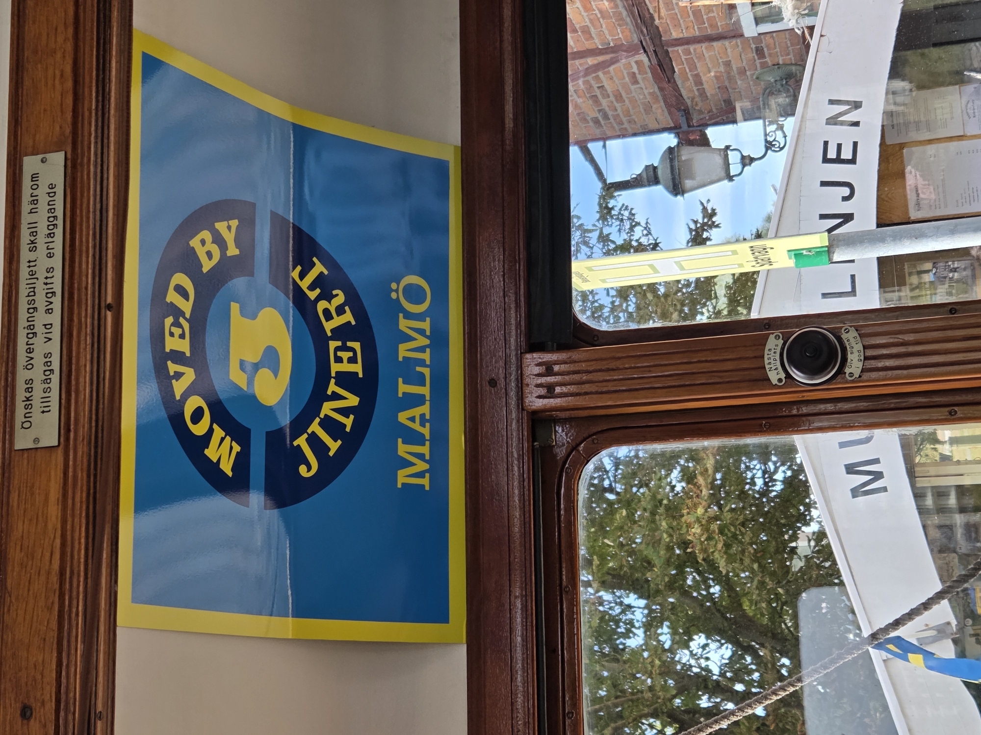This is an indoor, eye-level, slightly angled, close-up shot of a sign within a wooden-paneled interior, likely a tram or train car. The sign is predominantly blue and yellow, featuring the text "MOVED BY 3 JINERT MALMÖ" in yellow lettering arranged in a circular pattern. Below this, the word "MALMÖ" is written horizontally. A small, light-colored plaque with text is affixed to the wooden paneling above the sign, and a portion of the text is visible. Below the main sign, the interior of the vehicle continues, with wooden framing around a window. Through the window, trees and a brick building are visible, suggesting an outdoor environment. The text on the sign, "MALMÖ", indicates the city of Malmö, Sweden. There is a signal device with a button and text visible on the wooden panel to the right of the window. Further to the right, a green sign with "Spårvagn" is visible outside the window.