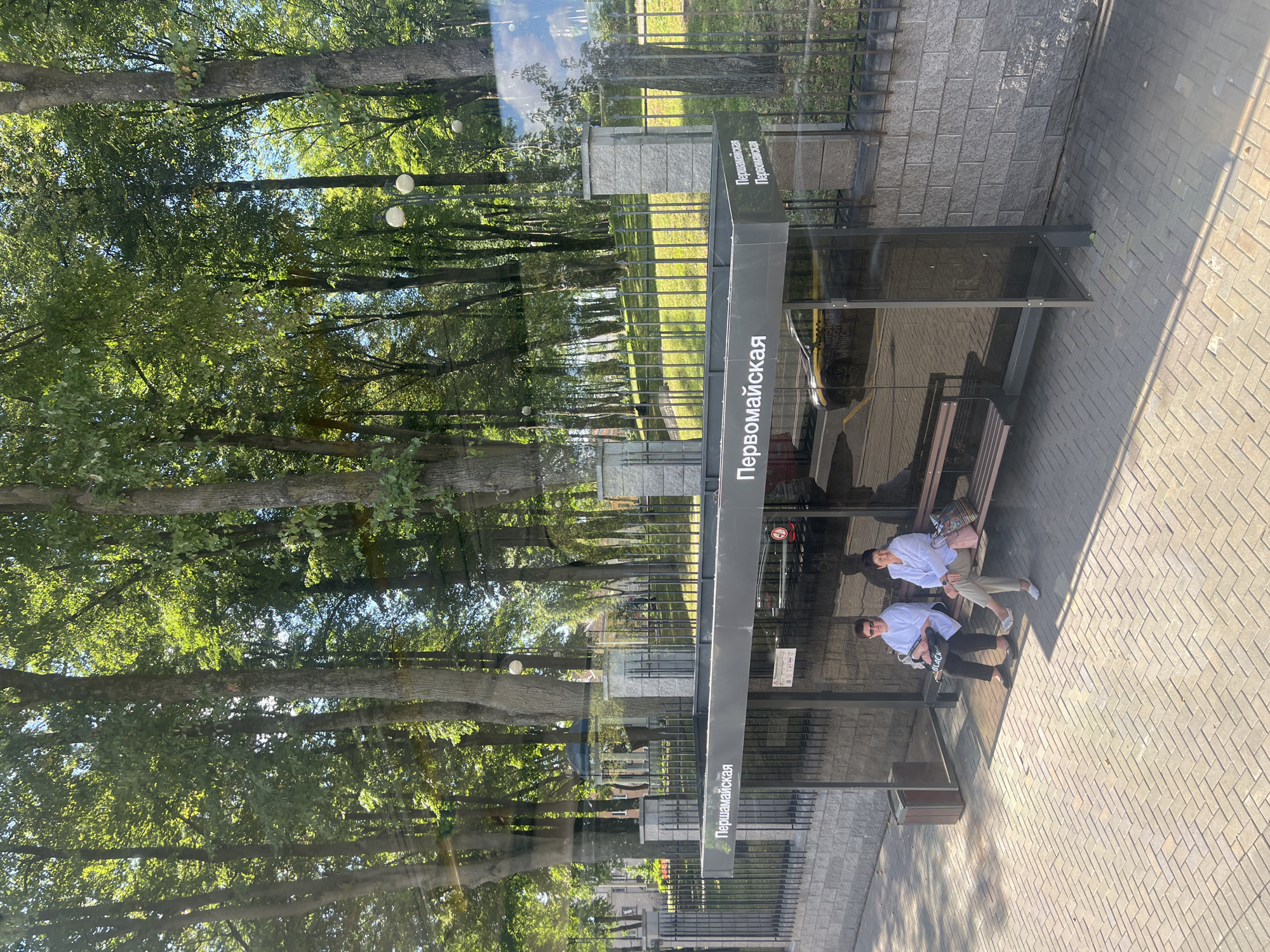 The image shows a bus stop shelter in Minsk, Belarus. The shelter has a dark gray roof and clear glass panels. The name of the bus stop, "Первомайская" (Pervomayskaya), is written in white Cyrillic letters on the roof. Two women are sitting on a wooden bench inside the shelter. Both women are wearing light-colored tops and dark pants. The woman on the left is wearing sunglasses and has a black bag with the word "BLACK" written on it. The woman on the right has a colorful patterned bag. Behind the bus stop is a black metal fence, a brick wall, and a grassy slope with trees. Sunlight filters through the trees, casting shadows on the pavement. A yellow taxi with black lettering is visible through the glass of the bus stop. The pavement is made of interlocking gray bricks.