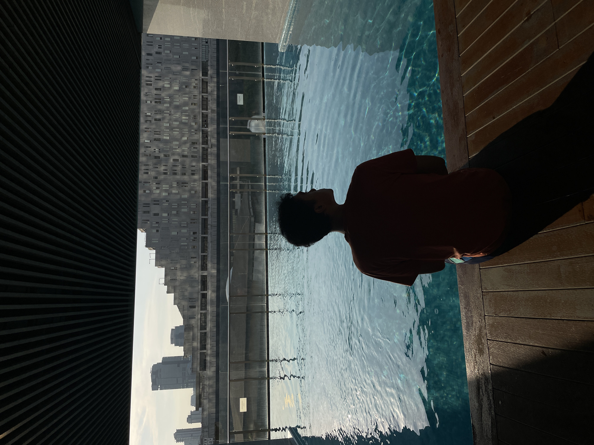 A person is sitting on the edge of a swimming pool in Central Jakarta, Indonesia. The pool is elevated, with a view of buildings and skyscrapers in the distance. The pool deck is made of dark wood planks. The pool is built at the top of a building. The photograph has a high angle view and has an unbalanced lighting.