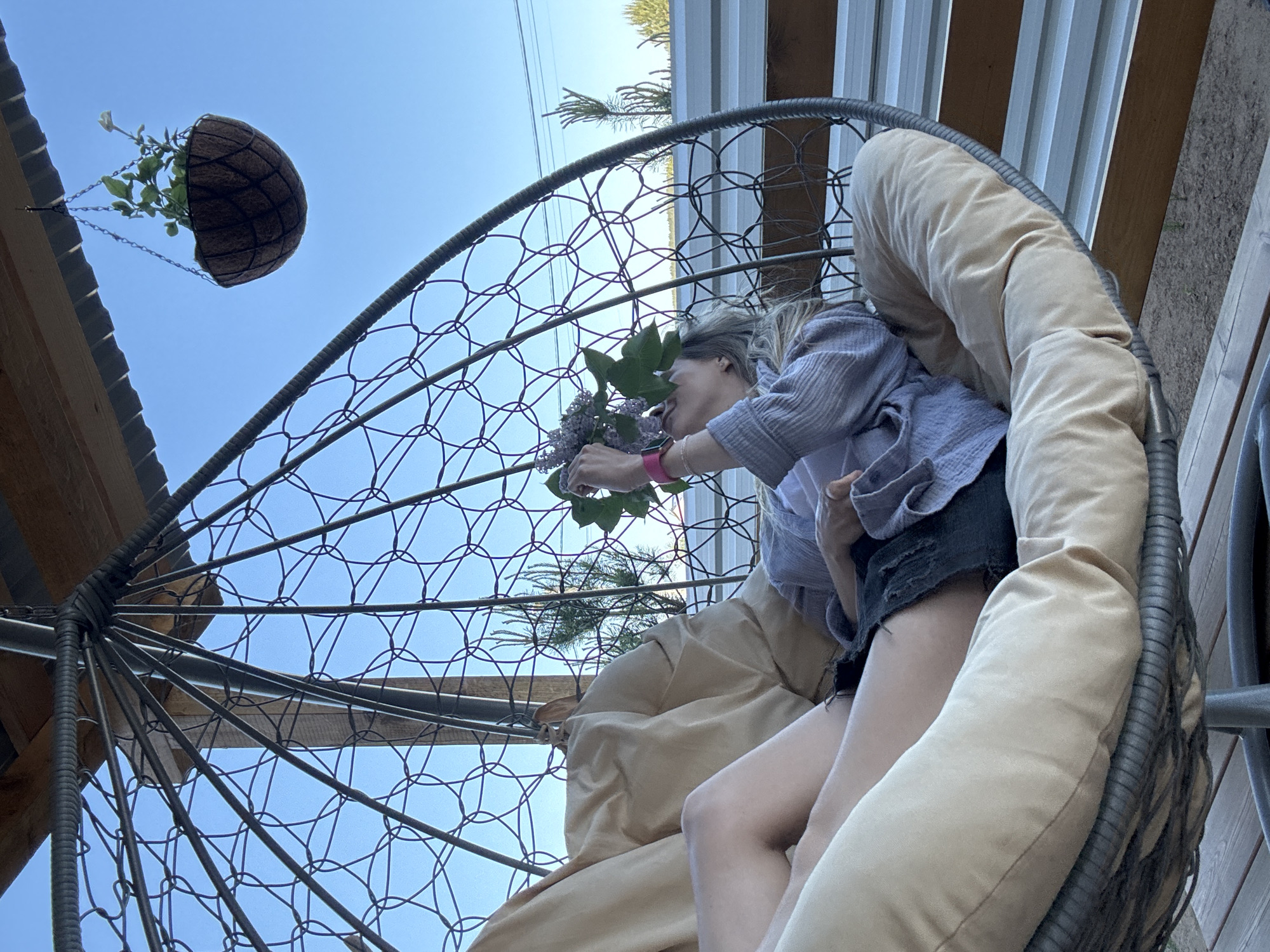 A woman is seated in a hanging chair outdoors, holding a bouquet of lilac flowers to her face. She wears a light purple button-up shirt over a white undershirt, dark shorts, and a pink watch. The chair has beige cushions and a wire frame. Above the chair, a hanging basket with green plants is visible. The sky is blue. Structural elements of a building are visible in the background and overhead. The location is Chaikovsky, Russia.