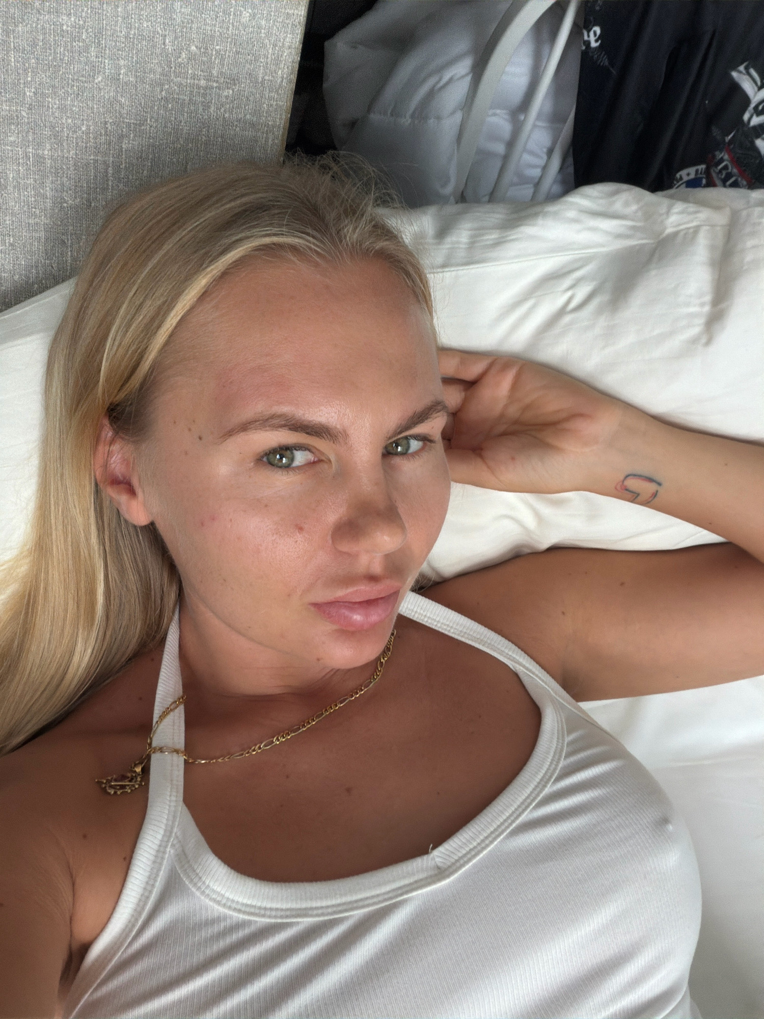 A blonde woman with green eyes is lying on a white pillow in bed. She is wearing a white tank top and a gold necklace. Her left arm is resting on her head, and she has a small tattoo on her wrist. The background shows a gray headboard and some clothing on a chair. The location is not specified in the city of Si Sunthon, Thailand.