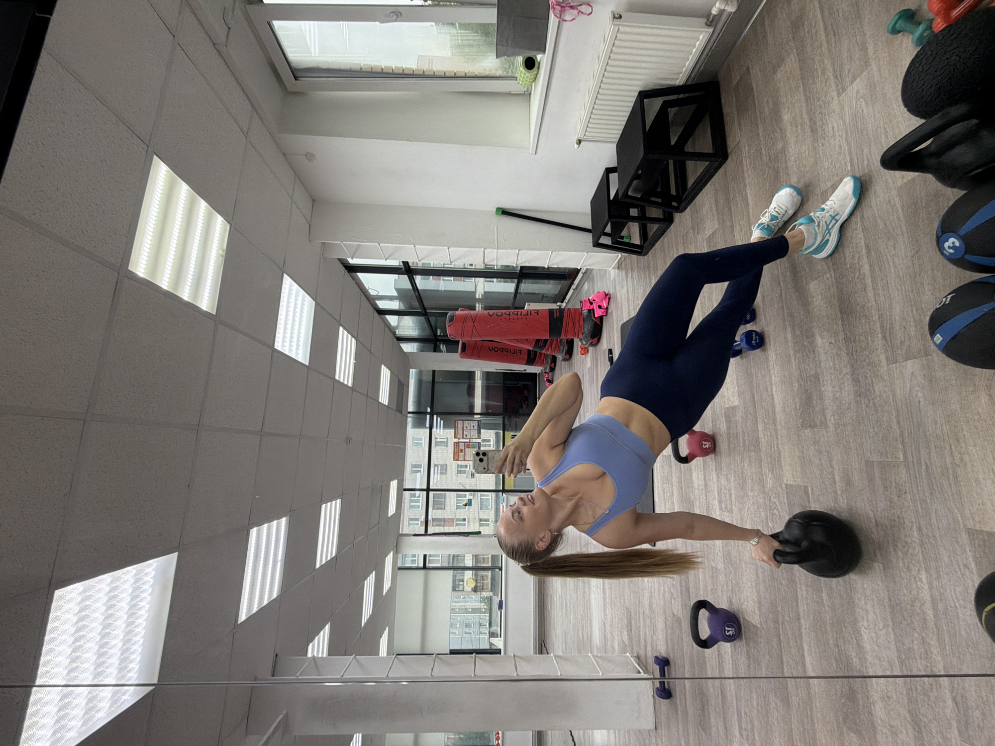 A woman in athletic wear performs a kettlebell exercise in a gym setting, holding a black kettlebell in her right hand. She wears a blue sports bra and dark blue leggings, and white athletic shoes with teal accents are on her feet. Her blond hair is in a ponytail. The gym environment includes visible ceiling lights, a window showing buildings outside (suggesting a city location), workout equipment, and a wooden floor. Based on the window view, this image was taken in Izhevsk, Russia.