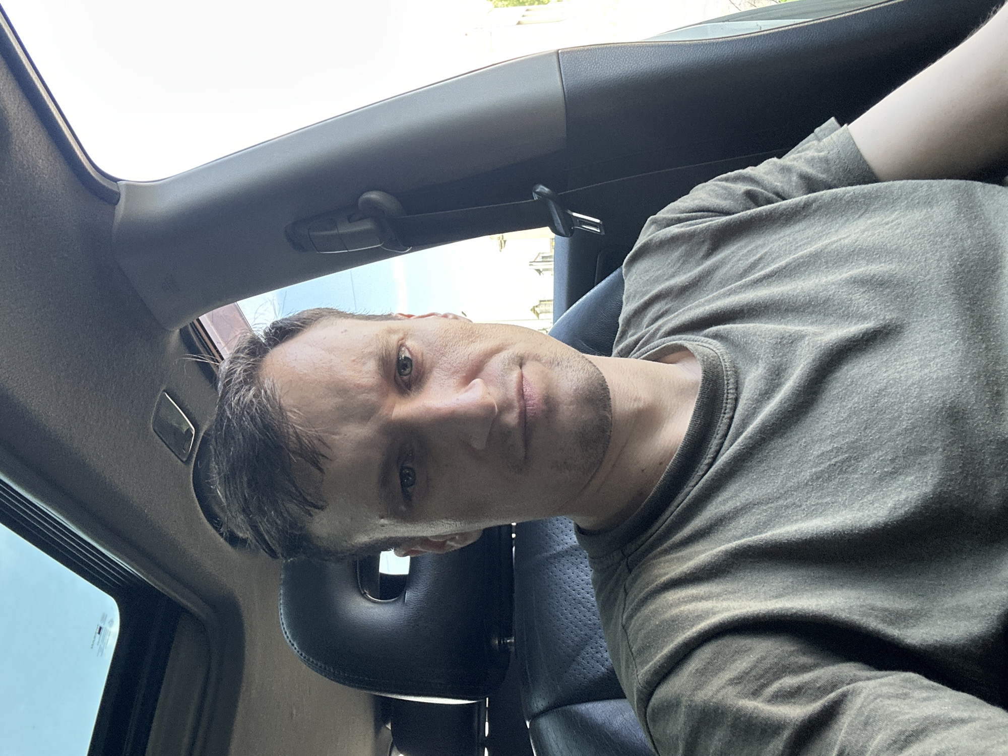 A medium shot portrait of a Caucasian male with short brown hair and stubble is captured from inside a car. He is wearing a dark green t-shirt. The car's interior is visible, including black leather seats, a sunroof, and a seatbelt buckle. The background suggests an outdoor, possibly urban environment, with a bright sky and hints of buildings. The specific location is identified as Shatava, Ukraine.
