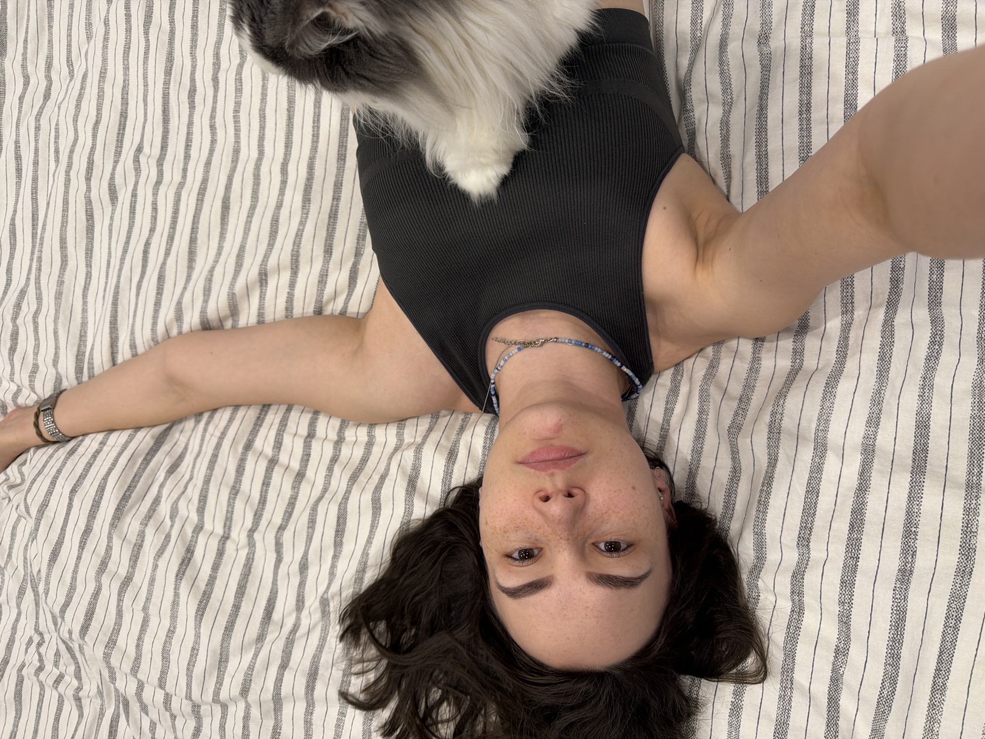 A person lies on their back on a striped bed, looking directly at the camera in a high-angle selfie taken in Chaikovsky, Russia. The individual, a young woman, has dark, curly hair spread around her head and a neutral to slightly contemplative expression, with visible freckles on her face. She is wearing a dark, ribbed tank top and a beaded necklace composed of blue, white, and silver beads. Her arms are extended to her sides, and a watch is visible on her left wrist (viewer's right). A fluffy cat, predominantly grey and white, is seen resting on her torso, with only its fur and a paw visible. The bed is covered with light-colored sheets featuring uneven, thin vertical grey stripes. The image captures a serene, indoor moment, possibly of relaxation or a casual interaction with a pet. No text is present in the media file.