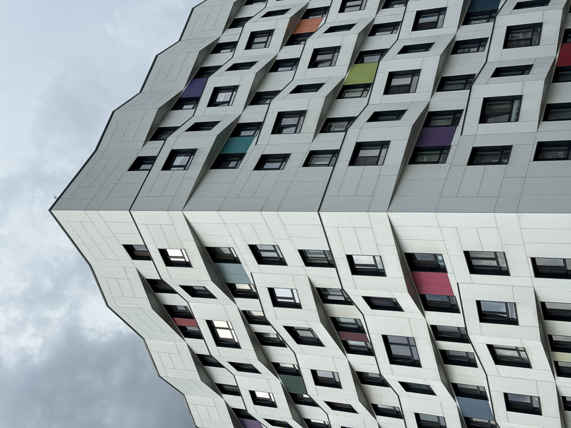 The image shows a modern building facade with a distinctive, geometrically complex design. The building is characterized by a repeating pattern of projecting and receding sections, creating a three-dimensional effect. Within these sections, square windows are uniformly spaced. Some window recesses have colored panels in shades of purple, orange, green, and blue. The building is likely located in Moscow, Russia, as indicated by the metadata. The sky is overcast. The lower half of the image is a reflection of the building.