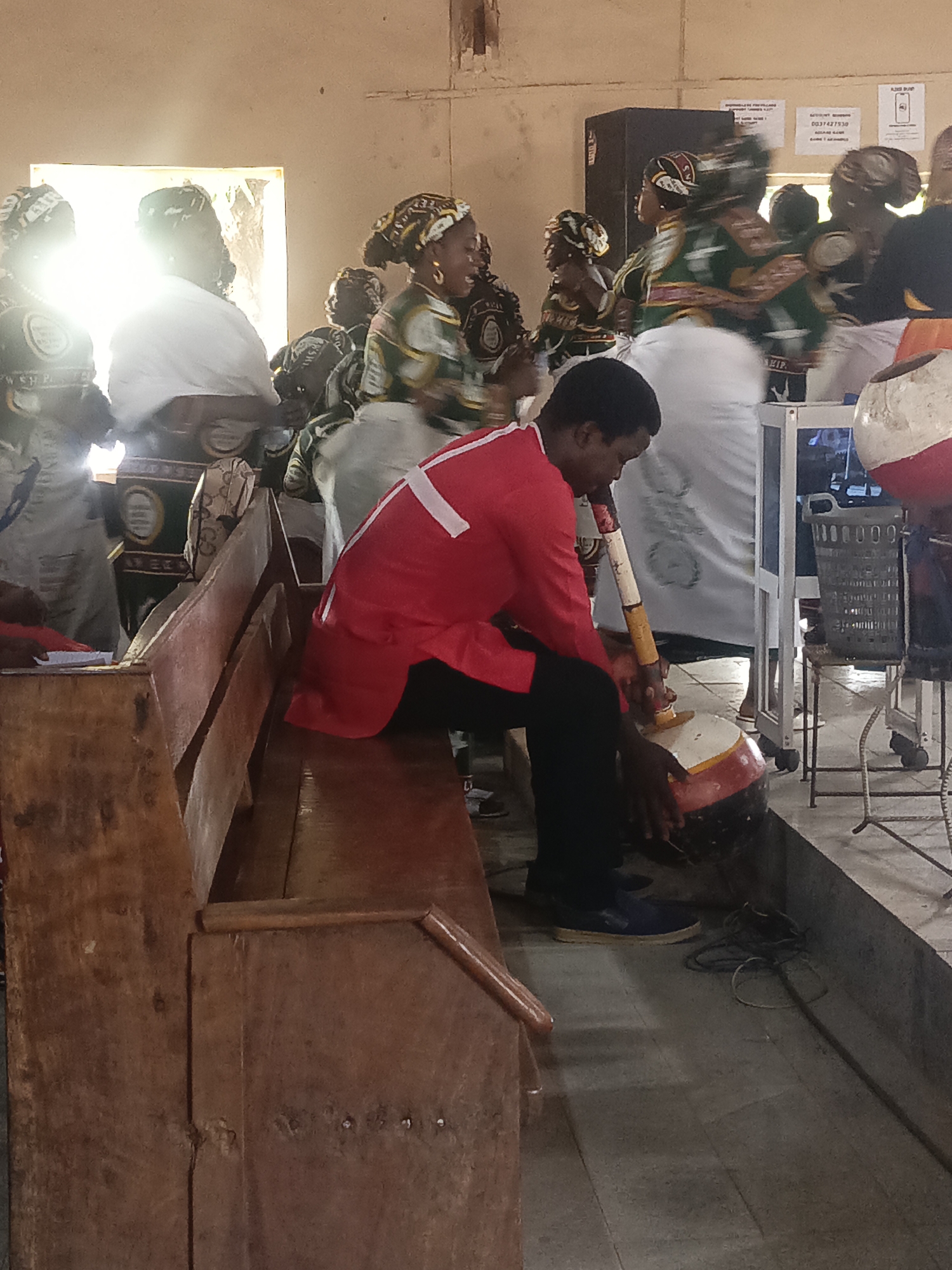 The image depicts an interior scene, likely in a church, in Akwanga, Nigeria. A man in a red shirt with a white cross on the back sits on a wooden bench playing a traditional drum. He is wearing black pants and blue shoes. Several women in traditional African dress are dancing or moving in the background. The background is somewhat blurred due to the movement of the women. Other people sit in the background. The light is bright. The environment appears to be simple and relatively unadorned. A trash basket is on a wheeled metal stand. There are cables on the floor.
