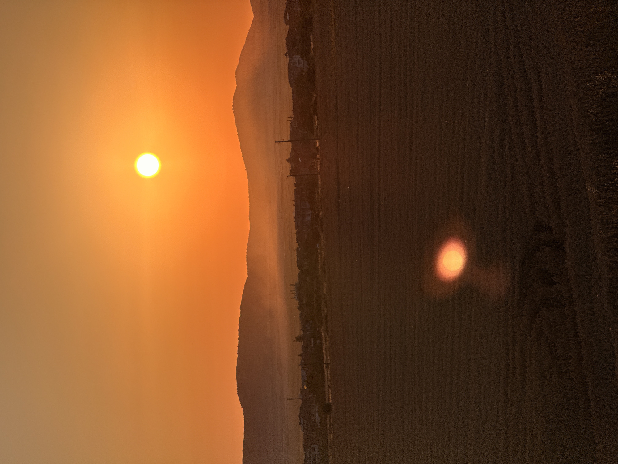 A wide shot captures a sunset over a rural landscape, likely near the city of Ravda, Bulgaria. The sky is a vibrant orange, transitioning to a lighter hue near the horizon. A bright, yellow sun is visible in the upper right quadrant of the image. In the midground, rolling hills are silhouetted against the sky, with a village nestled in the valley between them. The village appears to consist of low-rise buildings and some vegetation. In the foreground, a vast, dark field stretches across the lower half of the frame, with subtle textural patterns suggesting cultivation. A bright, elongated reflection of the sun is visible on the surface of this field. No people or animals are discernible in the image. The overall atmosphere is serene and depicts a rural sunset.