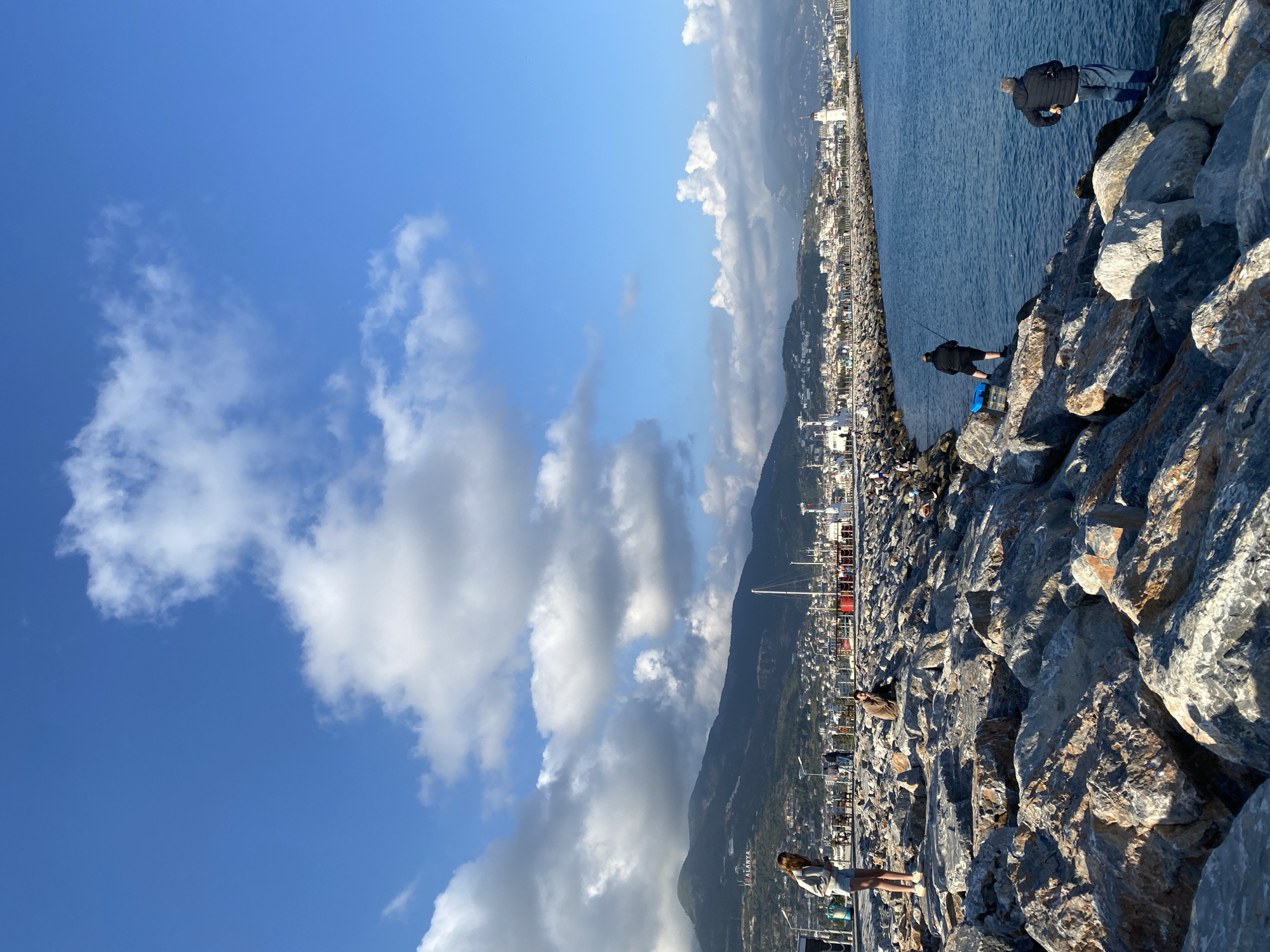 The image captures a vibrant coastal scene in Alanya, Turkey, on a sunny day, viewed from a rocky breakwater. The breakwater, composed of large grey and brown rocks, forms the foreground and extends diagonally from the bottom right towards the middle left of the frame.
Several individuals are visible on the breakwater. In the mid-ground on the right, two men are engaged in fishing from the rocks, one actively holding a fishing rod, with a blue cooler or box nearby. Closer to the camera, towards the bottom left, a woman in a light-colored top and shorts stands, appearing to observe the surroundings. Further along the breakwater towards the city, other people, including one wearing a brown jacket, are visible.
To the right of the breakwater, the calm, deep blue sea stretches horizontally, reflecting the bright sky. A white lighthouse-like structure is discernible in the distance along the breakwater. On the left side of the image, a bustling coastal city with numerous buildings sprawls along the shore, backed by large, verdant mountains. Faintly visible on the side of one mountain is the text "ALANYA," confirming the location.
The upper portion of the image features a clear, bright blue sky dotted with scattered white cumulus clouds. The strong sunlight casts visible shadows on the rocks and illuminates the entire scene, suggesting it is late morning or early afternoon. The overall atmosphere conveyed is one of leisure and activity, characteristic of a lively coastal area.