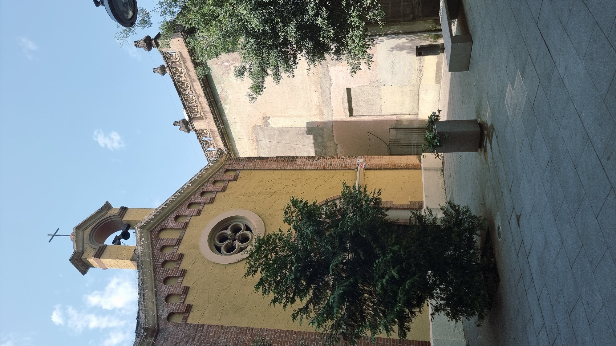 The image shows the facade of a building, likely a church, in Olot, Spain. The building features a yellow stucco wall with a textured pattern and red brick accents. A prominent feature is a rose window with a quatrefoil design. Above this, a bell tower with a cross is visible, housing a bell. Adjacent to the yellow building is a lighter-colored wall with visible signs of wear and patching. The foreground is a paved courtyard with dark gray rectangular tiles. A small evergreen tree stands to the left of the yellow building. Other elements in the courtyard include a tall, rectangular planter with greenery, a modern-looking gray bench, and a dark gray trash can. The sky is blue with scattered white clouds.