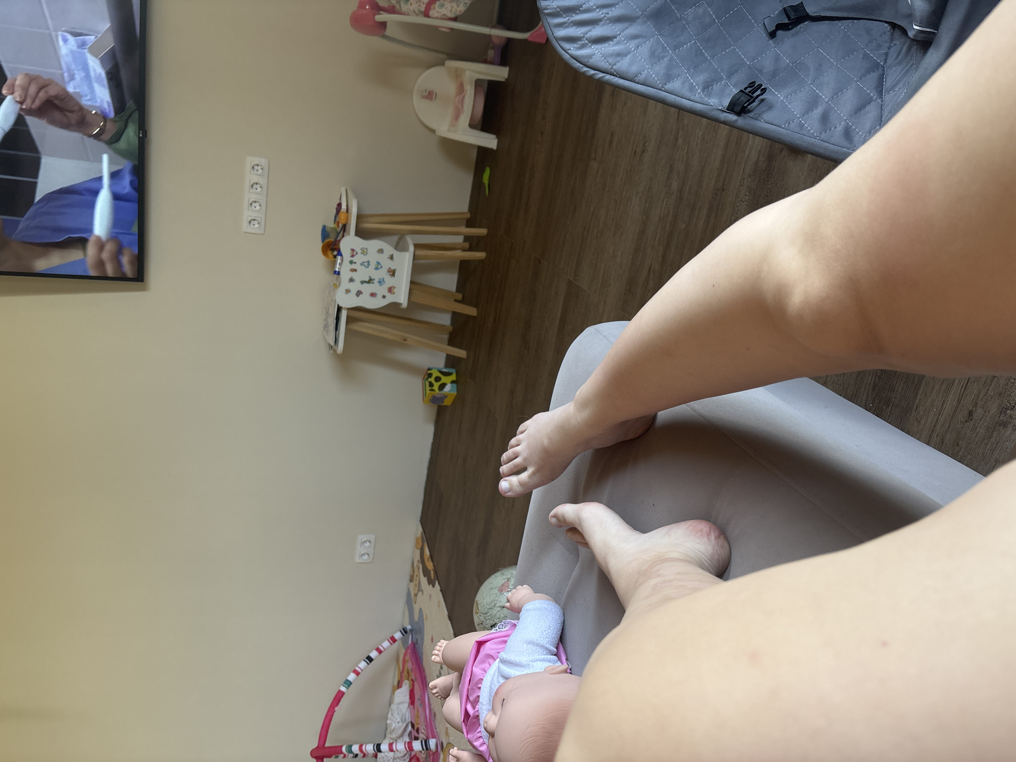The image shows an interior scene with a person's legs and feet visible, resting on a light gray surface, possibly a couch or chair. A doll wearing a pink dress and blue socks is positioned near the person's feet. In the background, there's a wall-mounted television displaying a scene involving hands and medical instruments. Further in the background, there's children's furniture, including a small table and chairs, and a play gym with colorful patterns. Electrical outlets are visible on the wall. Given that the location is Chaikovsky, Russia, according to the user, the image portrays a domestic setting, possibly a living room or playroom, with evidence of child-related activities and entertainment.