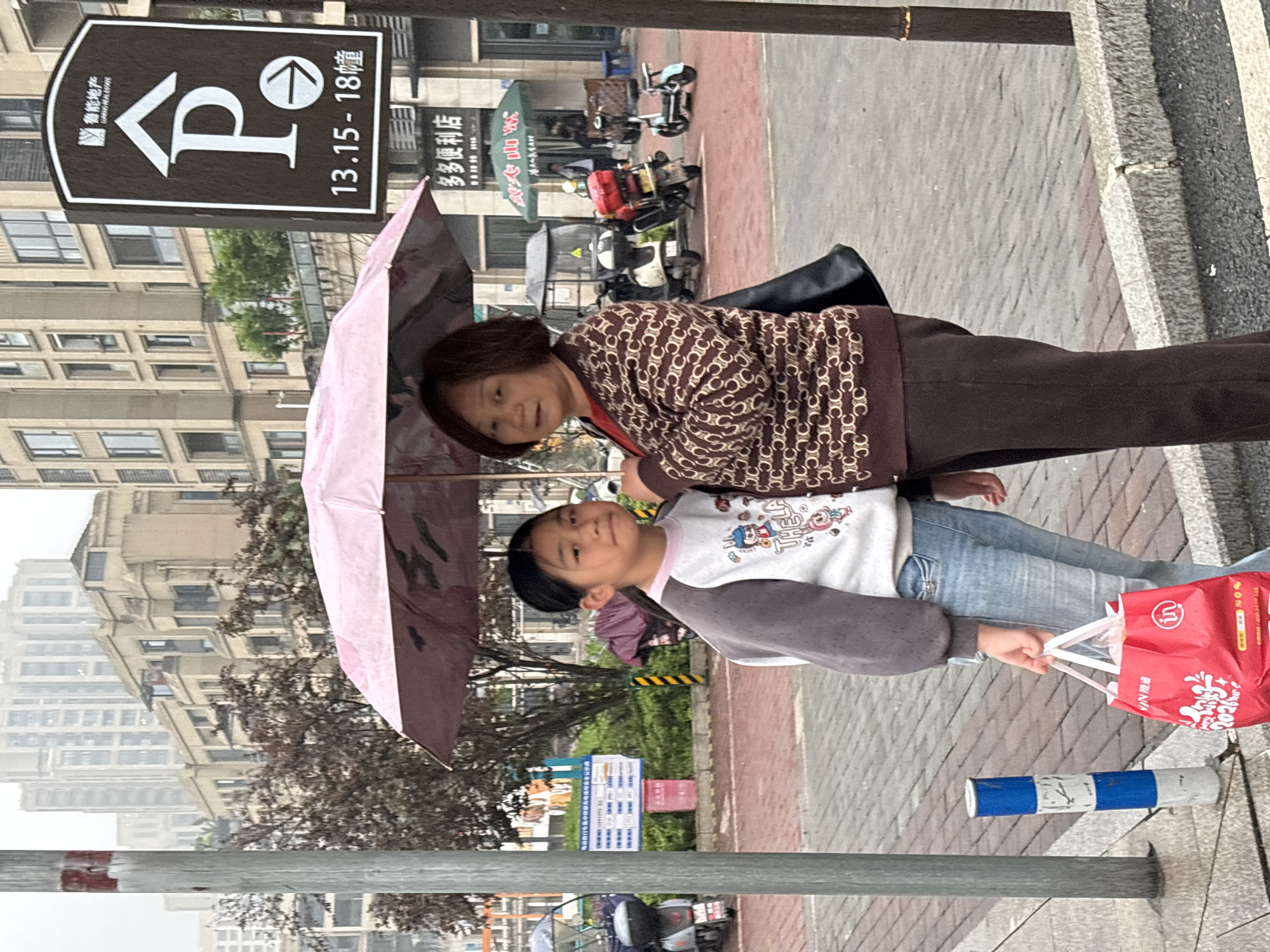 An eye-level medium shot depicts two individuals, likely a grandmother and granddaughter, walking on a paved sidewalk in what appears to be Jiangjin, China. The adult female is on the right, wearing a brown cardigan with a repetitive pattern and dark brown pants, and partially obscured by a light purple umbrella. The younger female, presumably her granddaughter, is on the left, dressed in a long-sleeved white shirt with cartoon graphics and light blue denim pants, holding a red shopping bag in her right hand. Both figures seem to be in motion, heading toward the right of the frame.
In the background, several buildings of varying heights can be observed, along with street signs featuring Chinese characters and numbers like "13.15-18". Parked motorcycles and other vehicles are also visible along the street. The environment seems to be urban, with wet pavement, possibly indicating recent rain. The overall composition suggests a mundane scene of daily life in a Chinese city.