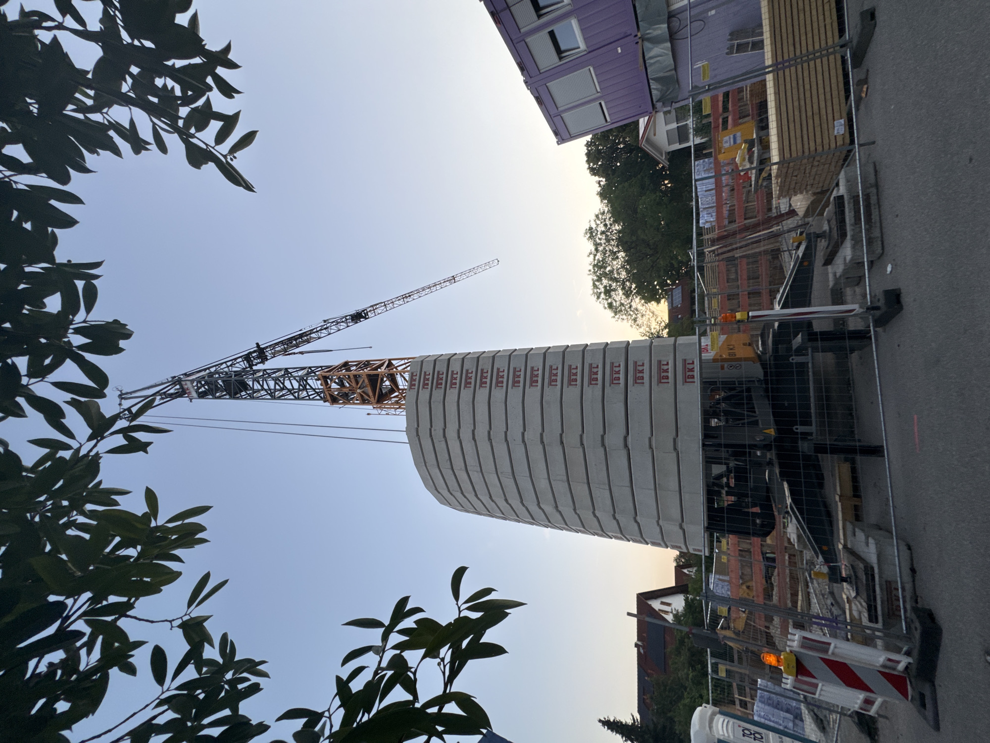 This is a photograph of a construction site at dusk in Höhenkirchen-Siegertsbrunn, Germany. The image is taken from a low angle, looking up towards the sky. In the foreground, dark green foliage of trees frames the scene. Dominating the center of the image is a large, curved structure made of stacked concrete segments, each marked with "IBKL".
A tall construction crane with a red and yellow jib extends into the sky, its arm angled downwards. Behind the concrete structure, a purple modular building with windows and closed shutters is visible, suggesting temporary accommodation or offices on site. Further back, a partially constructed building with wooden elements and red scaffolding can be seen. A chain-link fence encloses parts of the construction area, with traffic cones and barriers visible along the roadside.
The sky is a pale blue, transitioning to a warmer hue near the horizon, indicating the time of day is either sunrise or sunset. The overall atmosphere is that of an active construction project nearing the end of the day. Visible text on the concrete segments reads "IBKL".