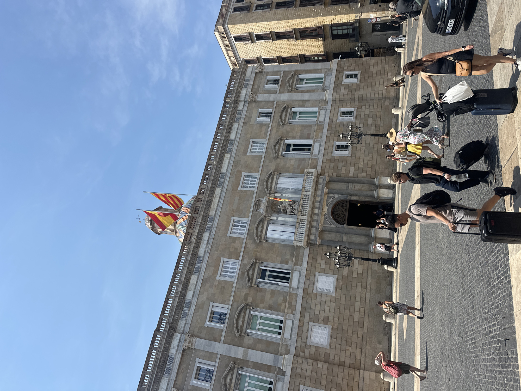 The image captures a sunny day in Barcelona, Spain, with a clear blue sky and scattered clouds. The scene is dominated by a grand, stone building with multiple stories, featuring numerous windows with white shutters, some open and some closed. Two flags, one red and yellow striped and another with yellow and red diagonal stripes, fly from a dome or turret atop the building. Decorative lampposts line the cobblestone plaza in front of the building.
Several people are present in the plaza, many of whom appear to be tourists. Some are walking, while others are with luggage, suggesting they are arriving or departing. One woman in a black dress is seen interacting with a stroller and luggage. Another individual, a man in a gray t-shirt and shorts, is pulling a black suitcase. A woman in a straw hat and a floral dress is also visible, along with other figures in the background, some entering or exiting the building. The shadows cast on the cobblestones indicate it is daytime, likely late morning or early afternoon. The atmosphere appears to be one of typical urban activity with a relaxed, travel-oriented feel. The building's architecture suggests a historical or governmental significance. Some text is visible on the building's facade, including "PALAU DE LA GENERALITAT" above an entrance.