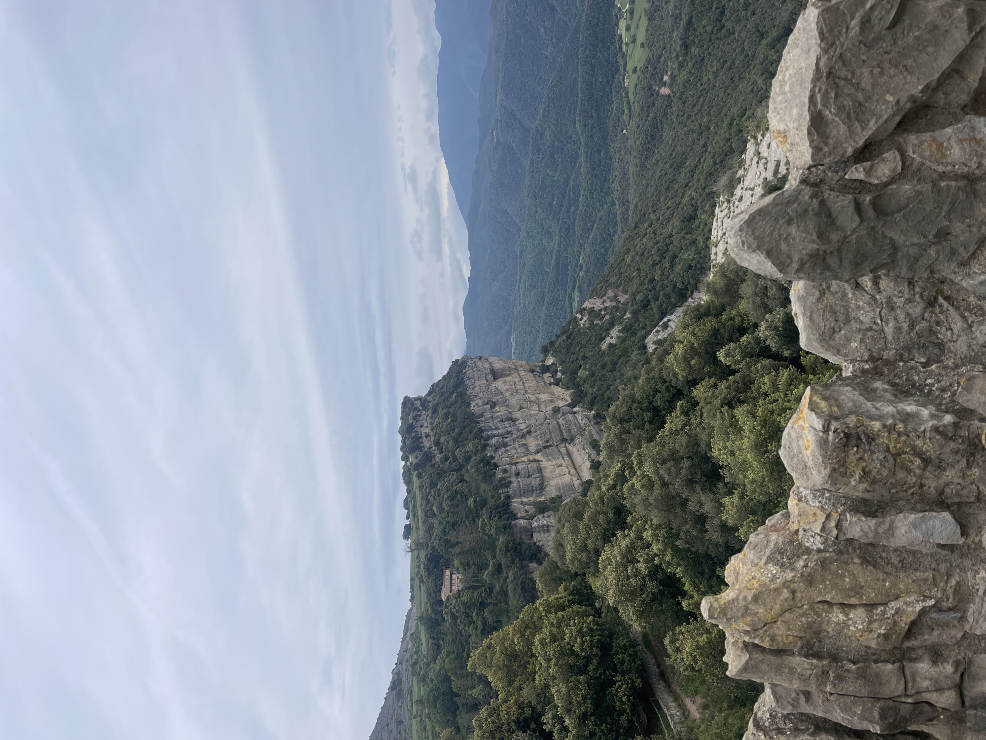 A landscape photograph depicts a mountainous terrain under an overcast sky, likely taken in Tavertet, Spain, based on the distinctive cliff features. In the foreground, rough, greyish-tan rocks form a natural barrier or overlook, partially obscuring the view. Beyond the rocks, a cliff face covered in dense green vegetation stands prominently. Further in the background, the landscape descends into a deep valley filled with similar lush greenery, fading into distant mountain ranges under a pale sky. The scene is serene, showcasing the natural beauty of the mountainous region. There are no people visible in the frame.