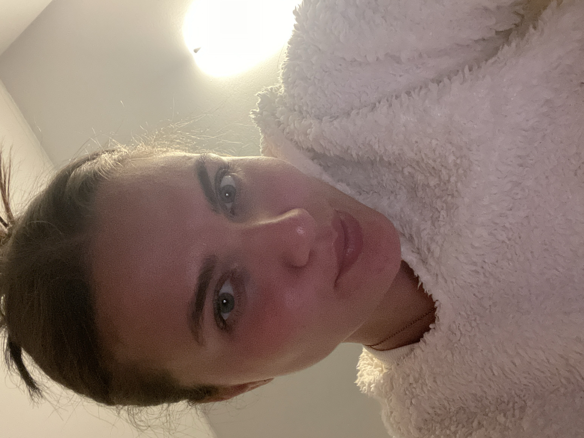A young woman with her hair pulled back is angled downward toward the camera. She has light skin, green eyes, and is wearing a soft, white, fluffy garment. A light fixture is visible in the upper background. The location of Мосметаллоконструкция Онт, Russia, is not determinable from the image.