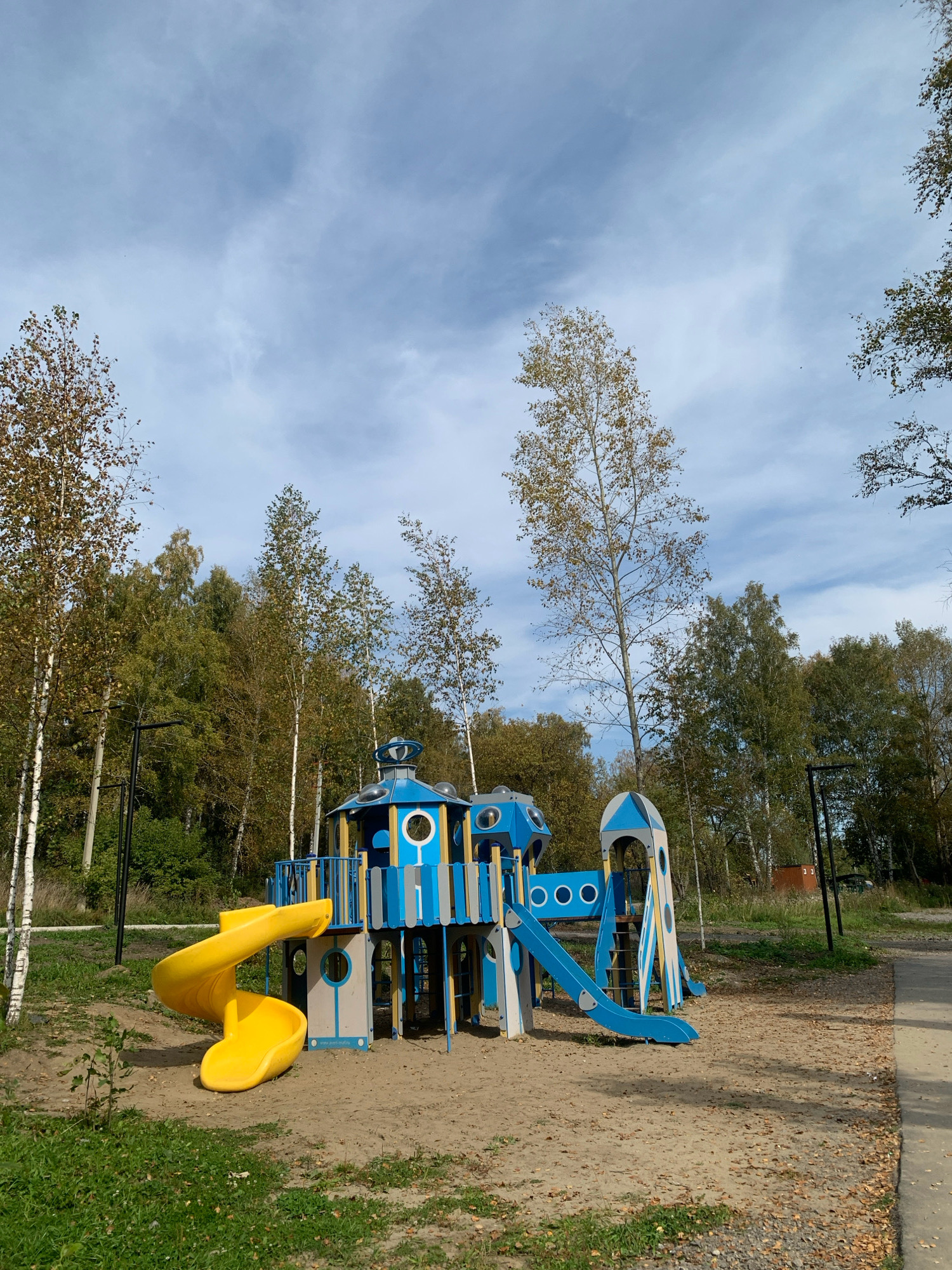 The image shows a children's playground located in Prokopyevsk, Russia, likely in a park or recreational area. The playground features a multi-colored structure with blue, grey, and yellow elements, including a yellow spiral slide, a blue straight slide, and various platforms, towers, and decorative circular openings. The playground is situated on a sandy surface surrounded by green grass and fallen leaves, indicating an autumn setting. In the background, a dense forest with deciduous trees, some showing autumn foliage, forms a natural backdrop. The sky is partly cloudy with a mix of blue sky and white, wispy clouds. Several tall, thin birch trees with white bark are visible in the foreground and middle ground, interspersed with the other trees. A black lamppost stands to the left of the playground. A paved path runs along the right side of the image. No people are present in the scene.