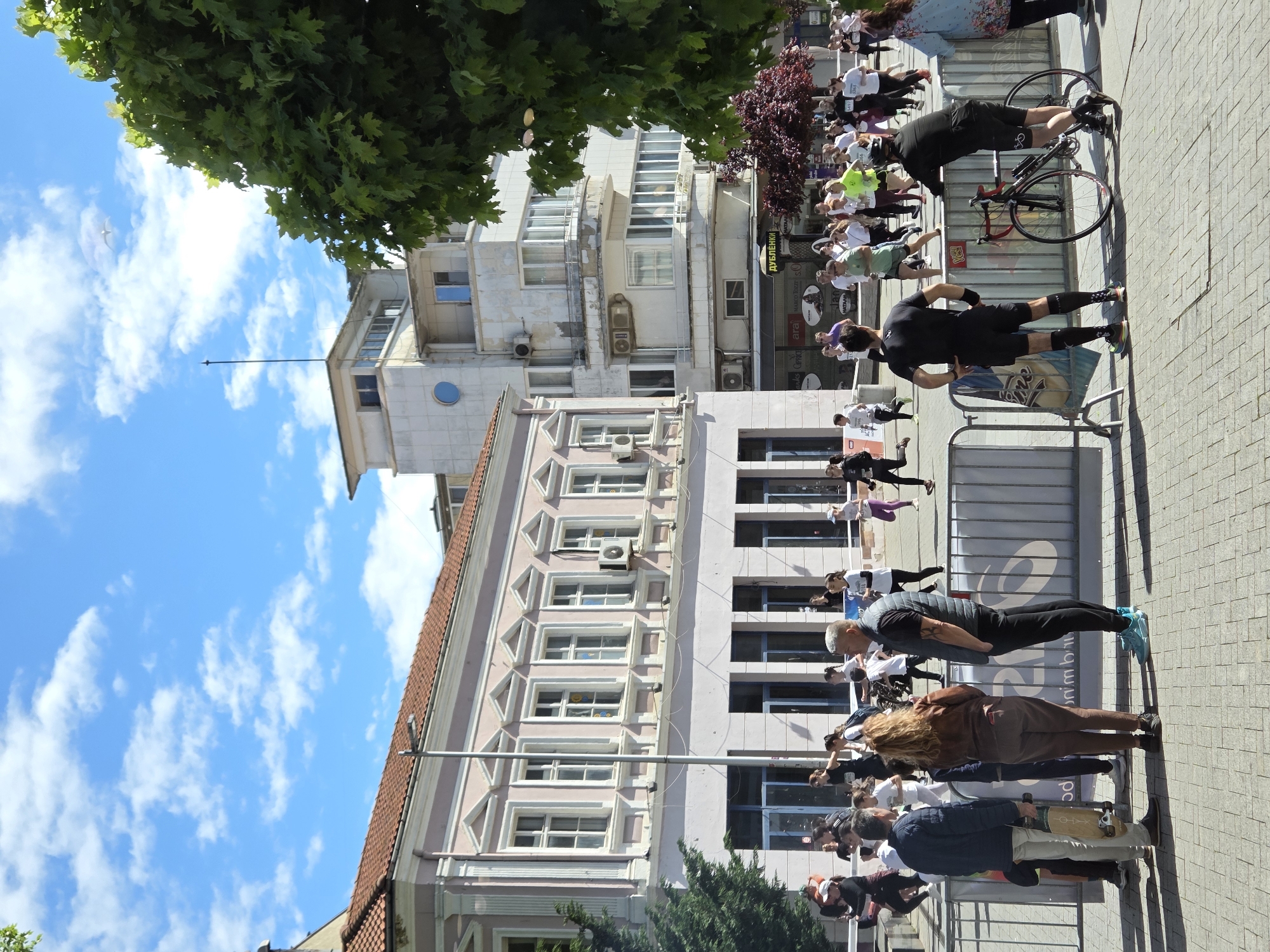The image shows a scene in Varna, Bulgaria. A group of people are participating in an event on a paved public space, likely a street or square. The event seems to be a race or walk, as the participants are moving in the same direction. A cyclist with a red bicycle is present among the participants.
Buildings form the backdrop, revealing the urban setting. The sky is blue with scattered clouds. Trees are visible, adding to the outdoor environment.