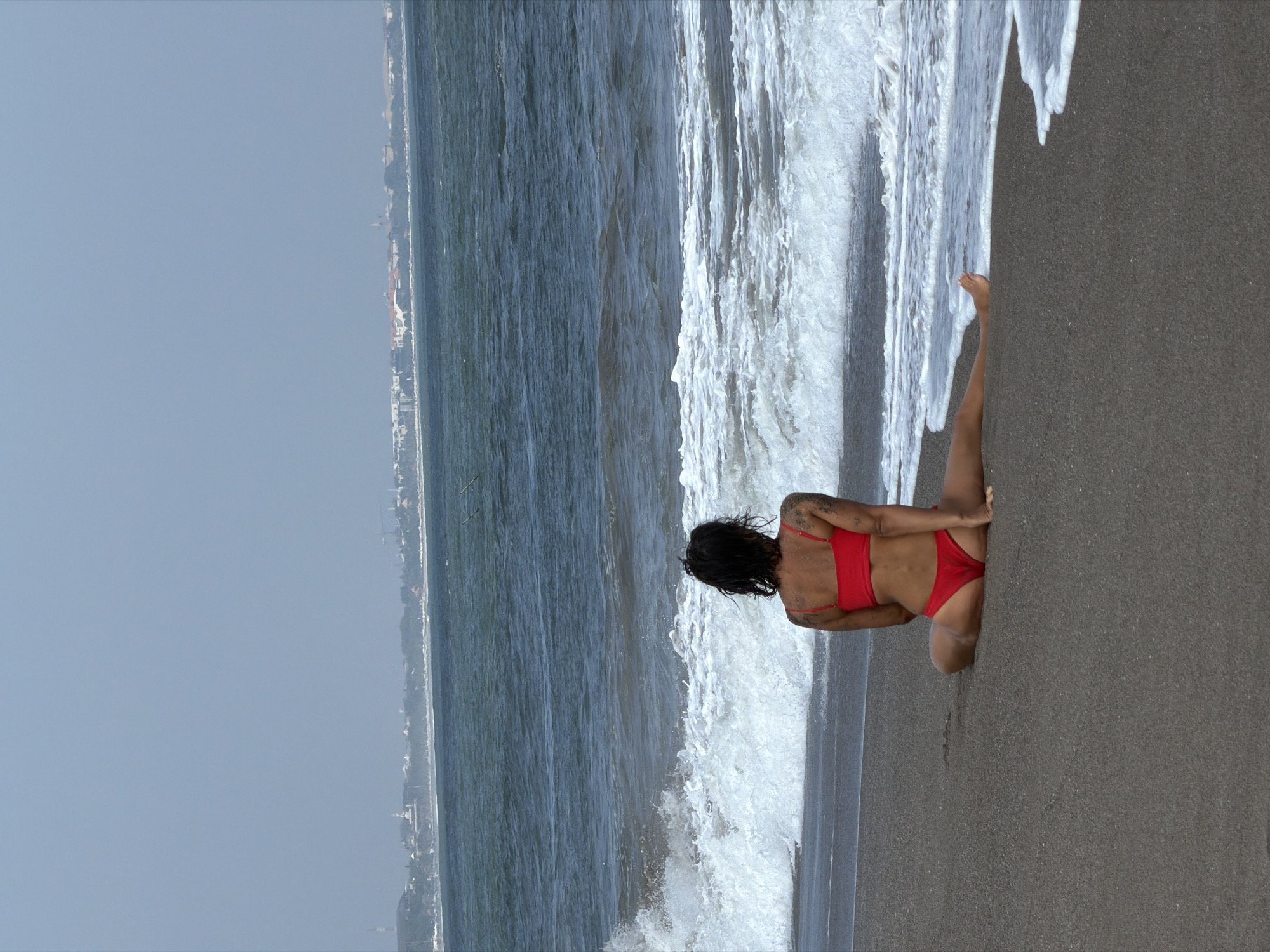 A woman with dark hair and tattoos on her arm is sitting on a dark sand beach, facing away from the camera. She is wearing a red bikini. The ocean is in front of her, with waves breaking on the shore. In the distance, a hazy city skyline is visible. The scene is likely located in North Kuta, Indonesia, a coastal area known for its beaches and tourist resorts.