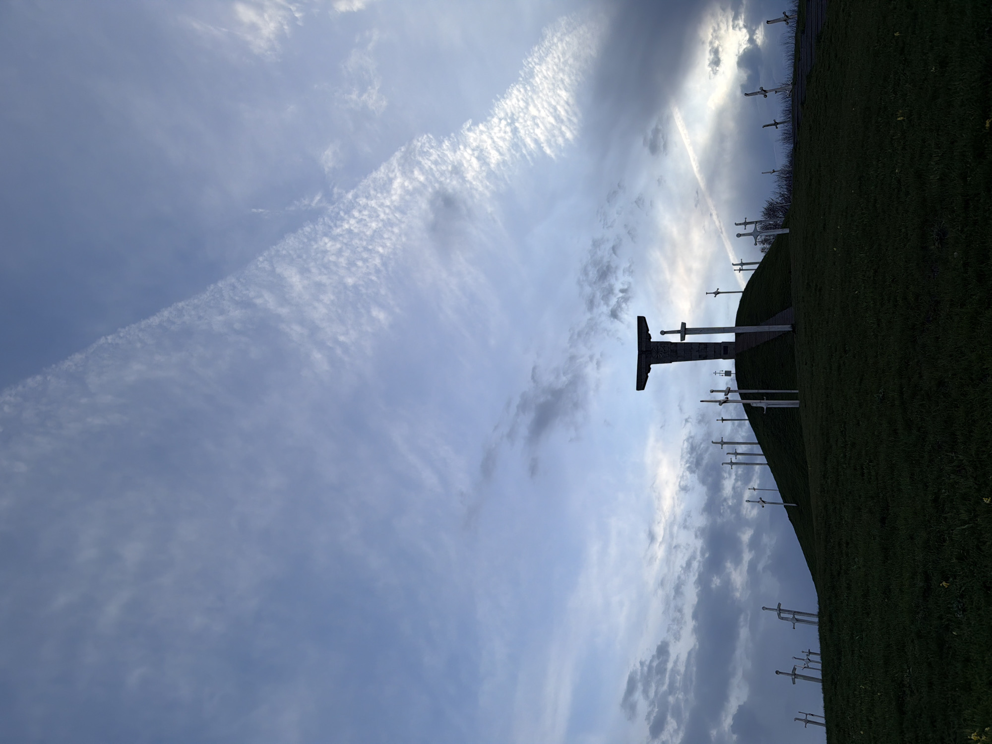 The image depicts a low-angle view of the Memorial of Georgian Soldiers in Zemo Nichbisi, Georgia. The sky is predominantly light blue with streaks of white clouds and a distinct contrail. A green hill rises from the lower right, dotted with numerous white sword-shaped monuments. At the crest of the hill, a large, dark sword-shaped monument stands out against the sky.