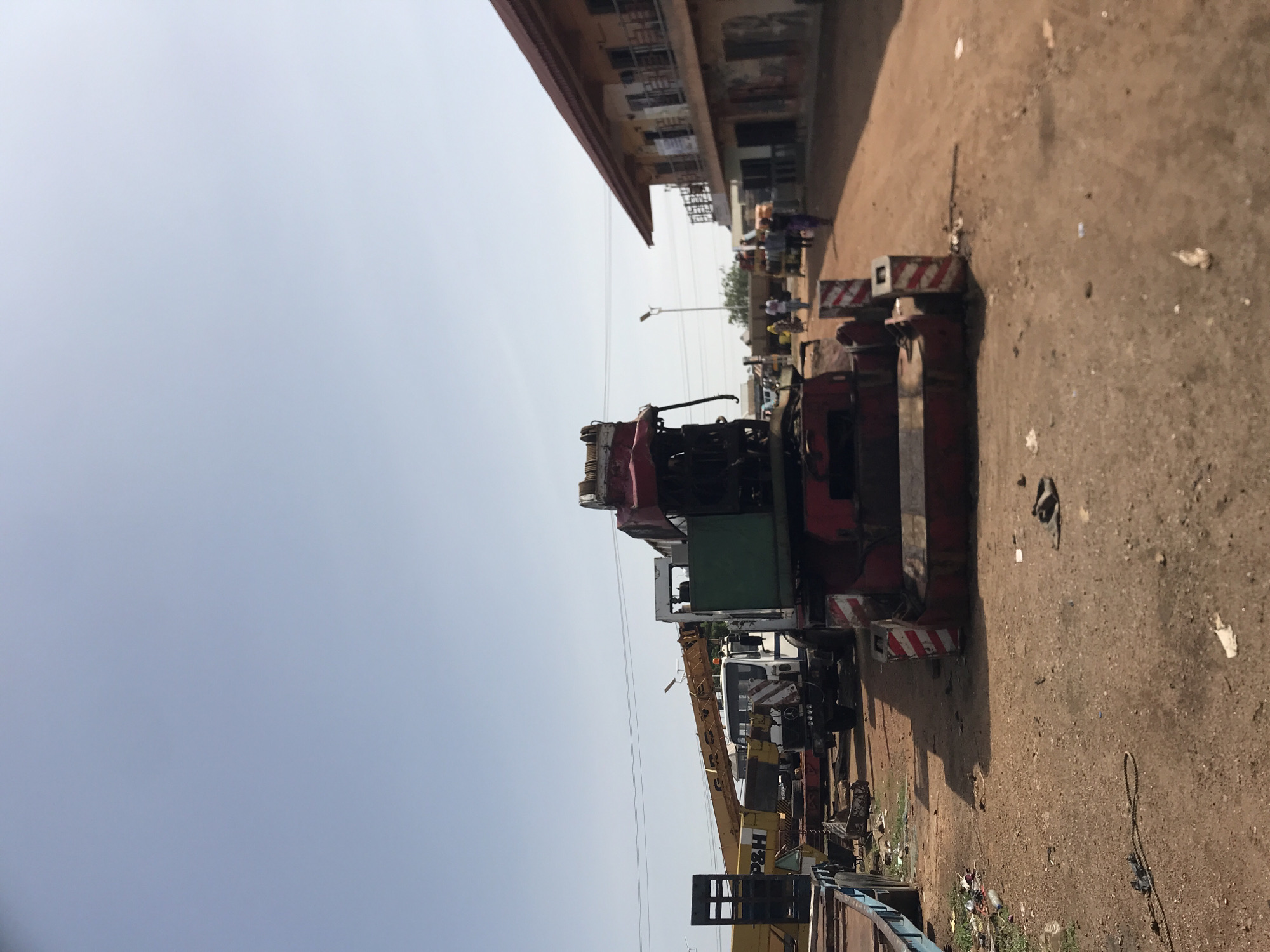 The image captures an outdoor scene, likely a street or industrial area in Jos, Nigeria, under a bright, clear sky. The photo is oriented vertically, with the sky on the left and the ground on the right.
In the foreground and midground, a line of heavy machinery, primarily cranes and trucks, is parked along an unpaved, dirt road. A prominent yellow crane with the text "P&H" visible on its side is seen among these vehicles. Other machinery parts are red and green, and some have red and white striped safety markings, possibly on outriggers. The ground is dusty and littered with various small debris, including what appears to be discarded plastic and wires.
On the opposite side of the road, to the left, stands a row of multi-story buildings. These buildings appear somewhat weathered, with visible windows and balconies. Several people are discernible in the distance along this side of the street, some standing, others possibly walking, but their features are indistinct. A tall street light pole is also visible in the midground, extending upwards towards the sky.
No specific active event is taking place; the machinery appears stationary, suggesting a parking or storage area. The clear, bright sky indicates it is daytime, likely morning or afternoon, with ample sunlight. The overall impression is that of a bustling, possibly commercial or industrial zone in an urban or peri-urban environment.