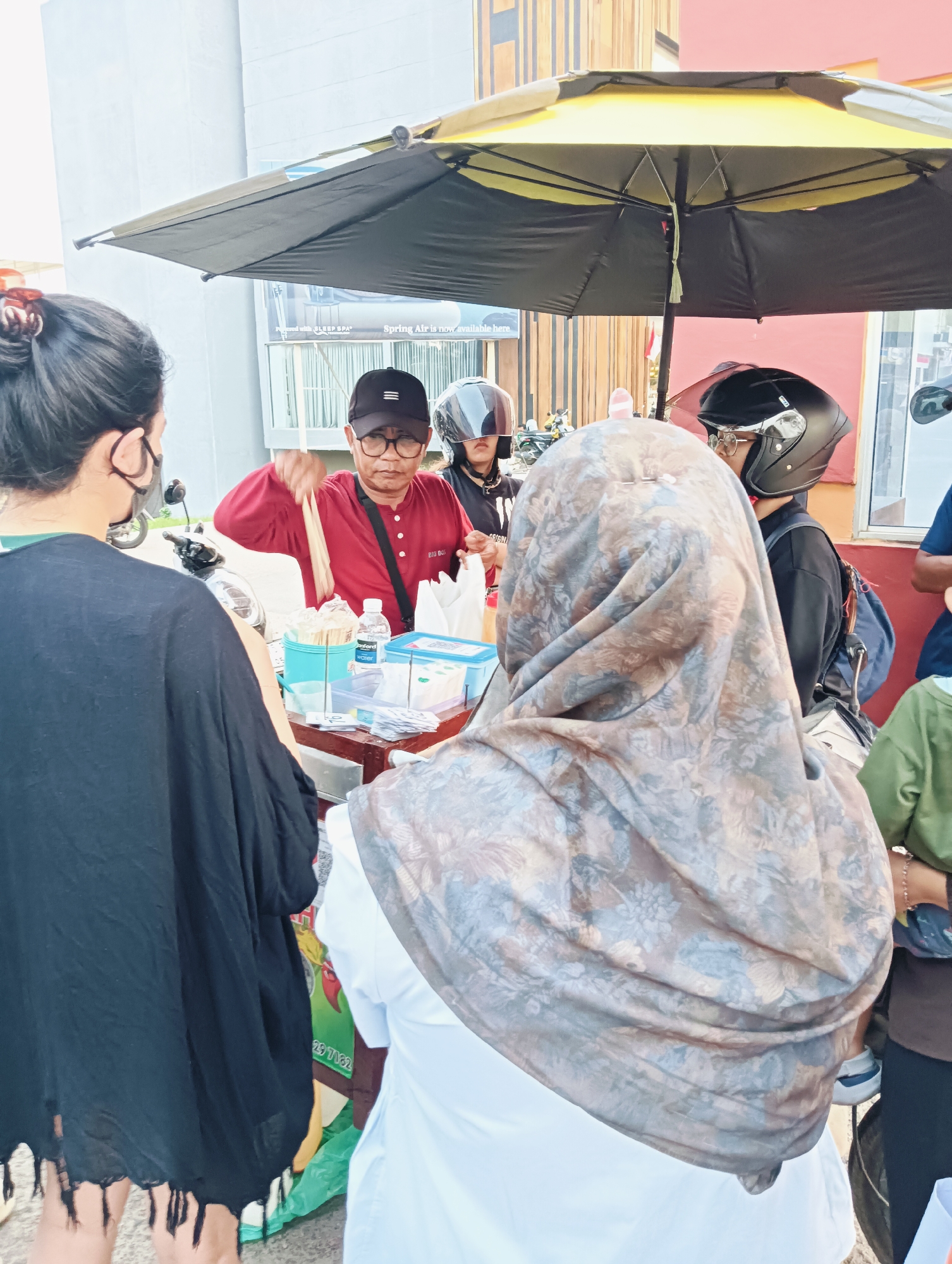 The image depicts a street food vendor in Batam, Indonesia, serving customers from a stall under a large umbrella. The vendor, a man wearing a red shirt and a black cap, is handing skewers to a woman wearing a black garment. Two other individuals, one wearing a grey hijab and the other a black helmet, are also visible near the stall, likely customers. The stall itself is cluttered with various items, including food containers, utensils, and bottled water. In the background, a building with an orange facade and windows can be seen, suggesting an urban environment. The overall scene conveys a sense of everyday commerce and daily life in a bustling city.