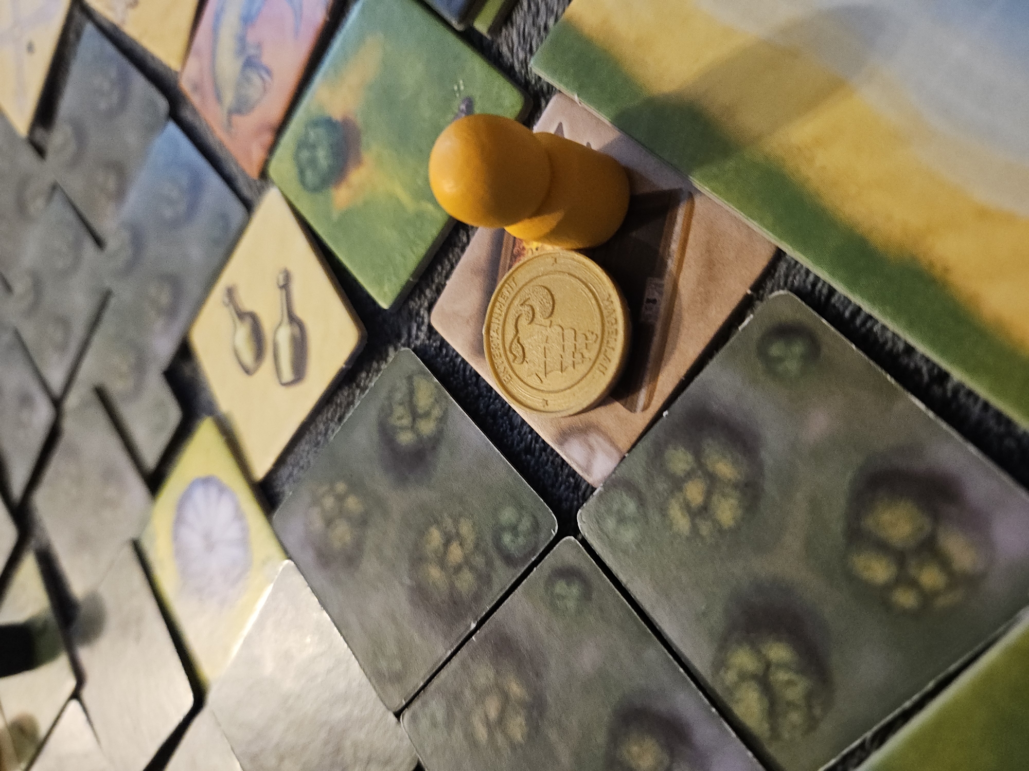 The image shows a collection of square game tiles arranged haphazardly. The tiles feature various illustrations, including hop plants, bottles, and other agricultural motifs. A gold-colored coin with an embossed figure and lettering is placed on top of one tile, and two orange-colored game pieces are stacked on top of the coin. The background is a dark, textured surface. The location is unspecified and unknown.