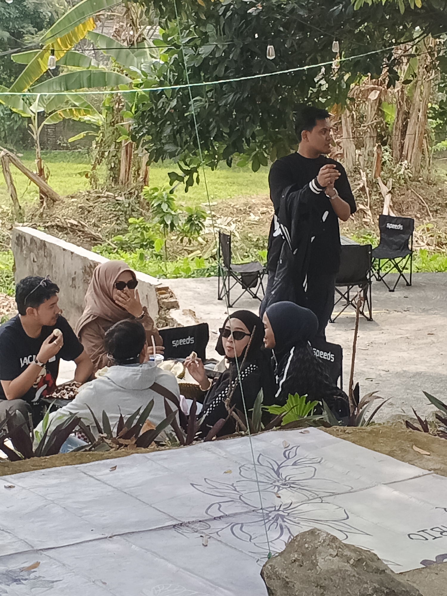 A group of six people are gathered outdoors, possibly at a cafe or rest stop in Pacet, Indonesia. The foreground features a large, patterned mat with floral designs, partially obscured by rocks and foliage. Behind this, several individuals are seated on the ground and on folding chairs.
To the left, a man in a black t-shirt with sunglasses on his head is eating. Next to him, a woman wearing a tan hijab and sunglasses is holding her hand to her mouth. In the center, a person in a light grey hooded jacket is facing away from the camera. To their right, two women are seated on black folding chairs, one wearing a black hijab and sunglasses, and the other with a dark blue hijab.
In the background, a man in a black t-shirt stands, holding what appears to be a dark jacket. Two black folding chairs with the "Speeds" logo are visible behind him. The environment suggests a semi-rural or natural setting with trees, foliage, and what looks like a dirt path or ground. String lights are suspended overhead, indicating a possibly decorative or functional lighting setup. The overall impression is a casual outdoor gathering.