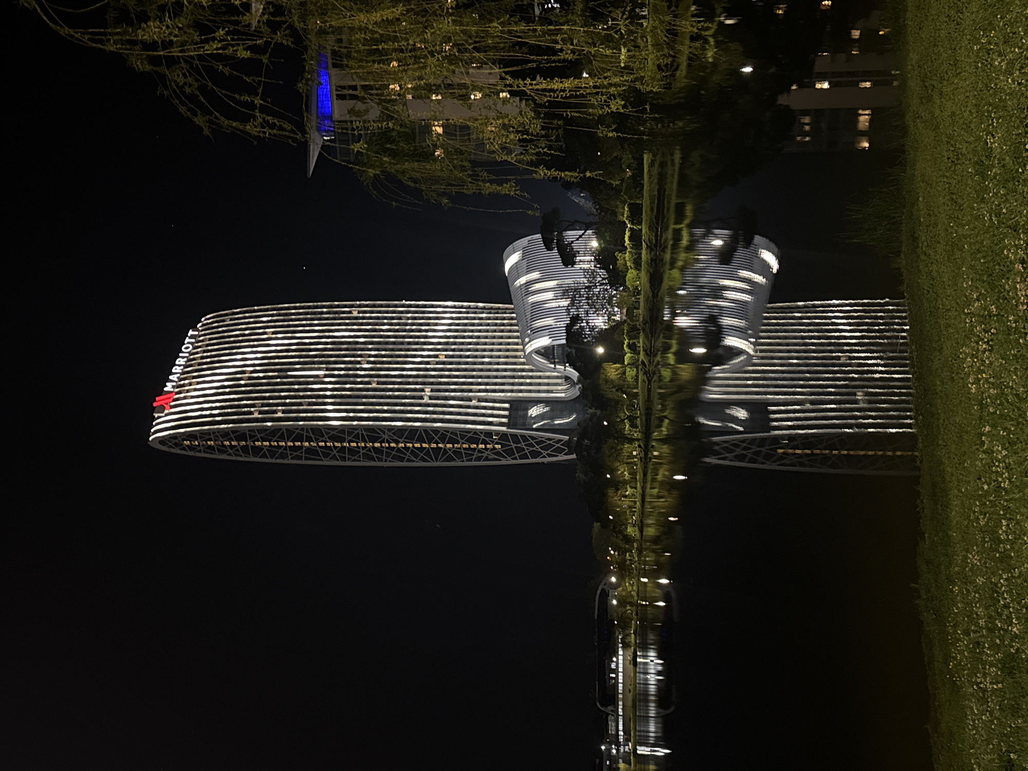 This nighttime image shows the Sheraton Hotel Batumi in Georgia, reflected in water. The hotel's horizontal lines of light are distinct, and a cylindrical structure is visible next to the main building. A tree and vegetation are present, partially obscuring the reflection. The environment suggests a dark, potentially urban setting.