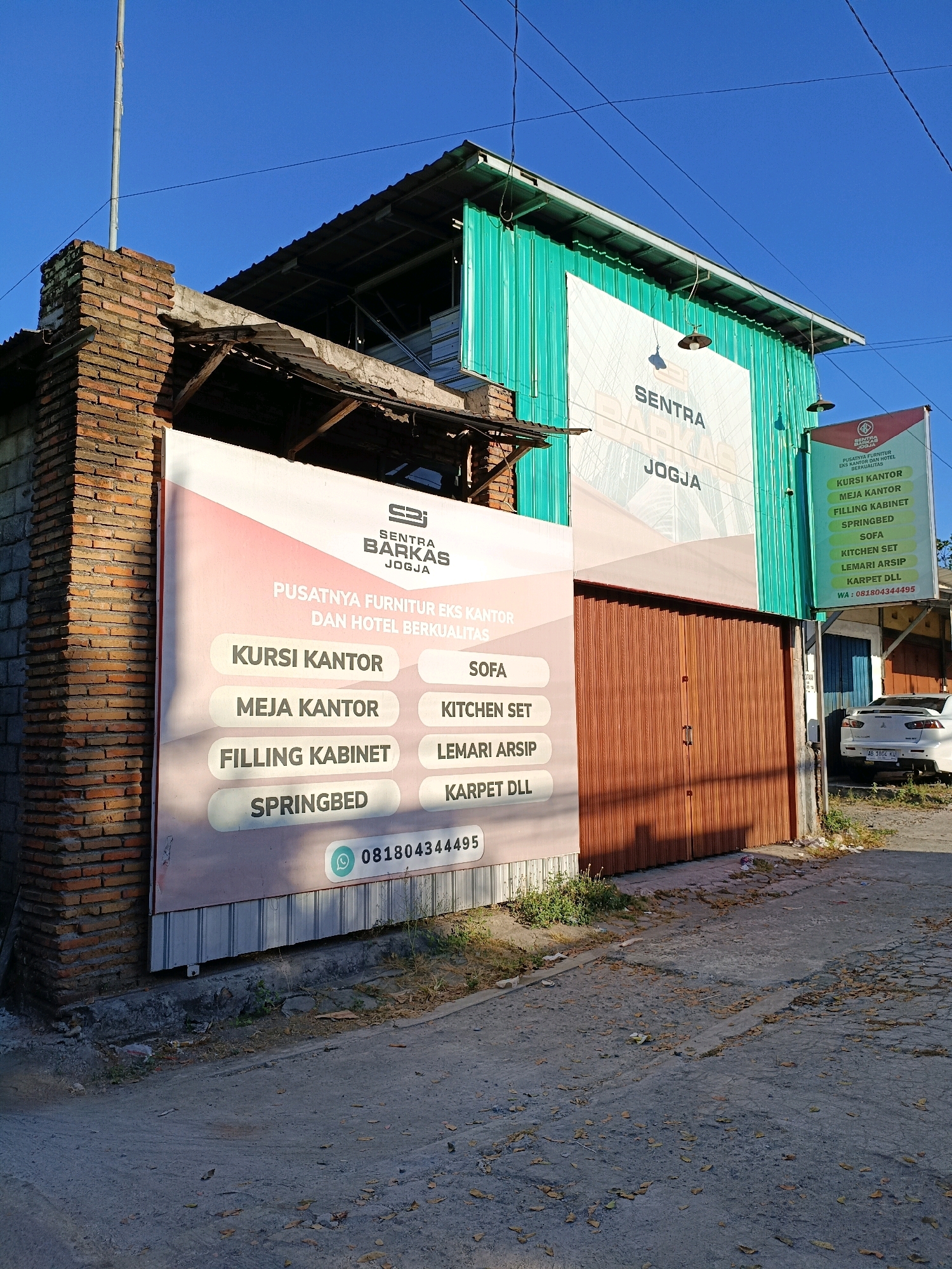 The image shows a building with a prominent sign in front of it, located in Maguwoharjo, Indonesia. The building appears to be a furniture store, as indicated by the text on the signs. One large sign, predominantly pink and white, lists various furniture items such as "KURSI KANTOR" (office chairs), "MEJA KANTOR" (office desks), "FILLING KABINET" (filing cabinets), and "SPRINGBED" (spring beds). It also displays a phone number, "081804344495". Another sign on the right side of the building, green and white, has similar text, including "KURSİ KANTOR", "MEJA KANTOR", "FILLING KABINET", "SPRINGBED", "SOFA", "KITCHEN SET", "LEMARI ARSIP" (archive cabinets), and "KARPET DLL" (carpets etc.), along with a phone number and "WA" indicating WhatsApp. Above these signs, the building itself is clad in green corrugated metal. The architecture suggests a commercial establishment, possibly with an attached workshop or storage area. A brick pillar stands to the left of the main sign. A white car is parked to the right of the building. The ground in front of the building is paved with asphalt, showing signs of wear and littered with fallen leaves. The sky above is clear and blue.